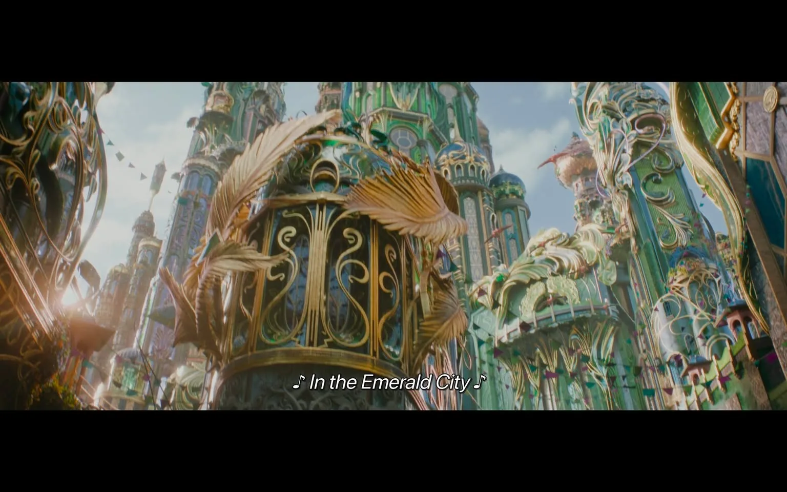 In the image, a stunning view of a fantastical city is presented, characterized by intricate architectural designs and vibrant colors. The buildings are adorned with elaborate decorations, including golden elements and flowing, organic shapes that resemble leaves and feathers. The sky is bright, suggesting daytime, and there are colorful banners strung between the structures, adding to the festive atmosphere. The text at the bottom indicates a musical element, with the phrase "In the Emerald City" suggesting a connection to a narrative or performance. Overall, the scene conveys a sense of wonder and enchantment typical of a magical setting.