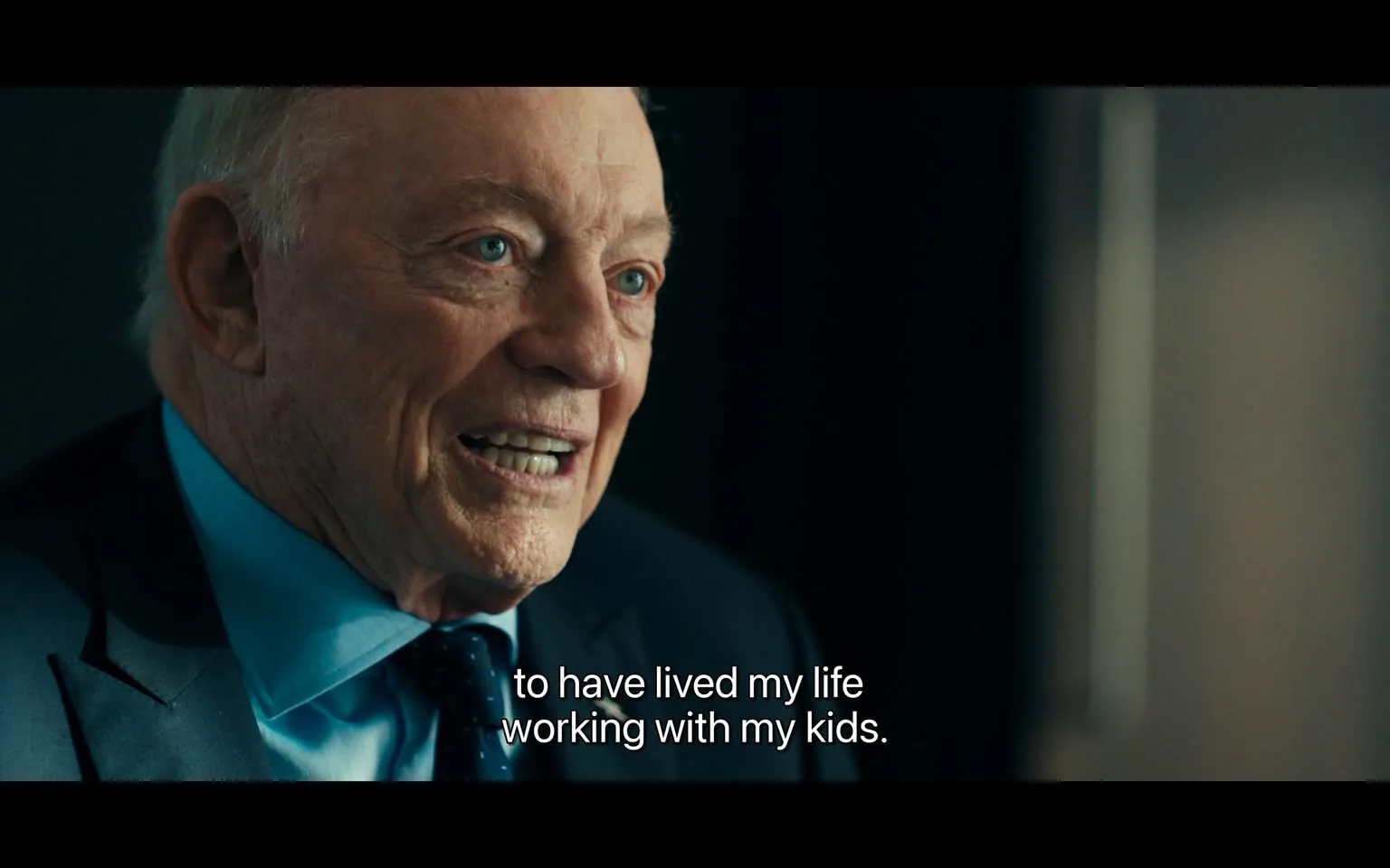 In the image, an unidentified adult male is engaged in conversation, displaying a warm smile and expressive eyes. He is dressed in a dark suit with a light blue shirt and a patterned tie. The background appears to be a softly lit indoor setting, possibly an office or interview space, with a blurred backdrop that suggests a professional environment. The visible text conveys a personal sentiment about cherishing time spent with family, specifically mentioning working with his kids. His expression suggests a sense of fulfillment and nostalgia as he reflects on his experiences.