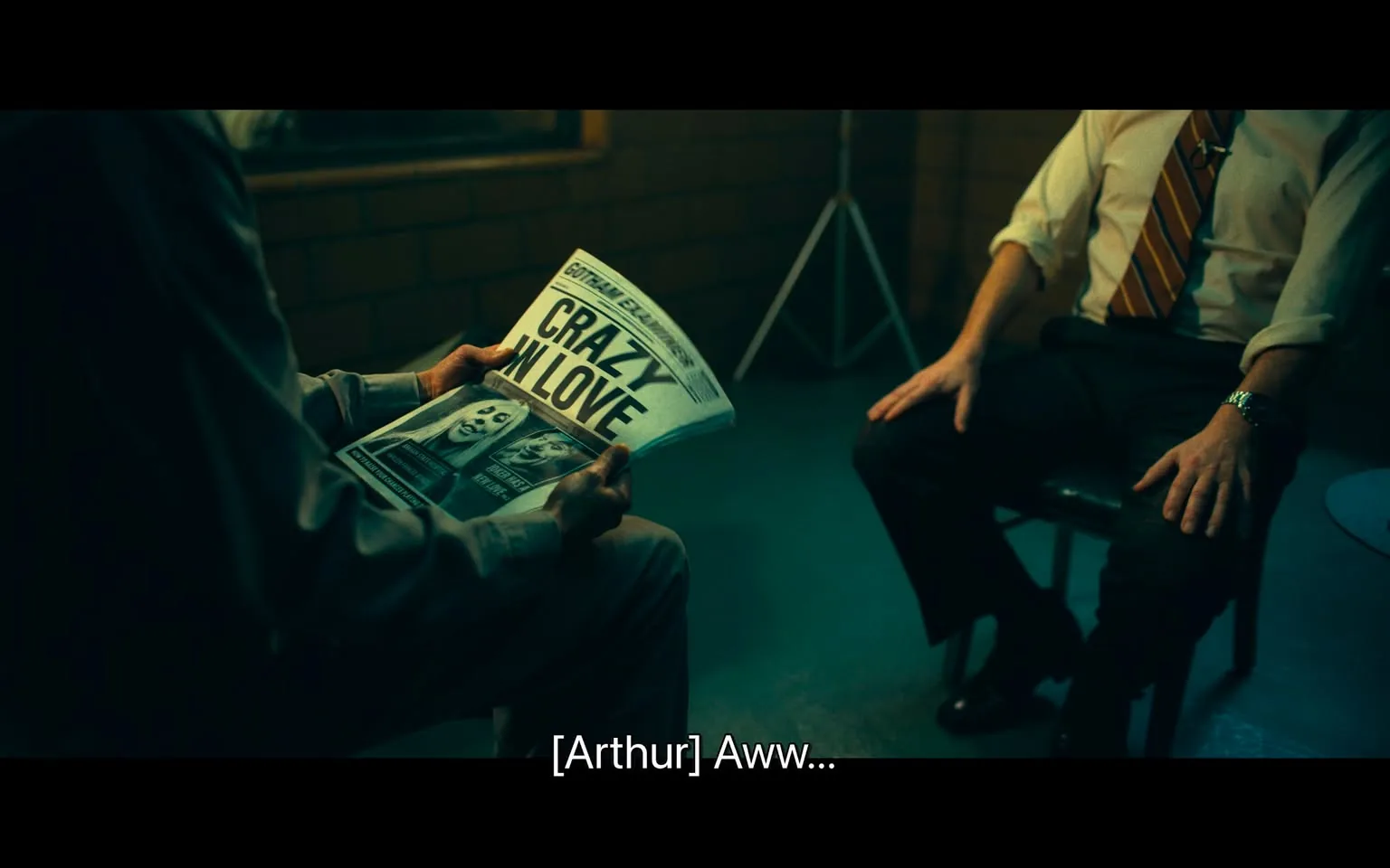 In a dimly lit room, two individuals are seated across from each other. One person, dressed in a light-colored shirt, is holding a newspaper titled "CRAZY IN LOVE," which features images of individuals with painted faces, likely suggesting a connection to a character or theme. The other person, wearing a white shirt with a striped tie, sits attentively, hands resting on their knees. The setting appears to be an interrogation or interview space, indicated by the sparse furnishings and the presence of a tripod in the background. The atmosphere is tense, with the person holding the newspaper expressing a sentiment of "Aww..." suggesting an emotional reaction to the content.