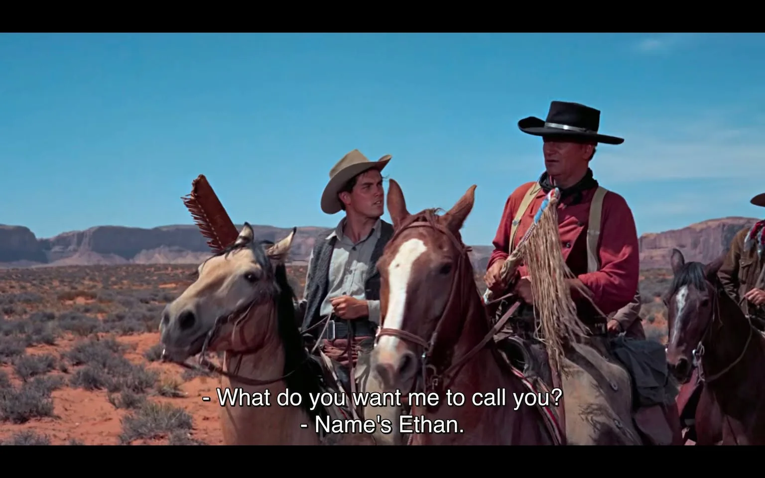 In a desert landscape, two individuals are riding horses and engaged in a conversation. One rider, wearing a light-colored cowboy hat and a button-up shirt, appears to be younger, while the other, dressed in a red shirt and a black hat, seems older and more authoritative. The younger rider looks inquisitive, while the older one introduces himself as Ethan. The background features rocky formations and a clear blue sky, indicating an outdoor setting typical of a western film. The horses are well-groomed, with one displaying a distinctive mane. The dialogue suggests a developing relationship or partnership between the two characters.
