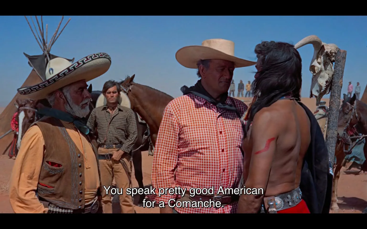In the scene, two men are engaged in a confrontational dialogue, with one character making a remark about the other's ability to speak English. The man on the left has a long, white beard and wears a large sombrero, while the man in the center is dressed in a checkered shirt and a black scarf. The third man, standing to the right, has long hair and is shirtless, displaying a red mark on his arm. The background features horses and a desert landscape, with a few figures visible in the distance. The setting suggests a Western theme, likely in a rural or frontier environment. The dialogue hints at cultural tensions, reflecting historical interactions between different groups.