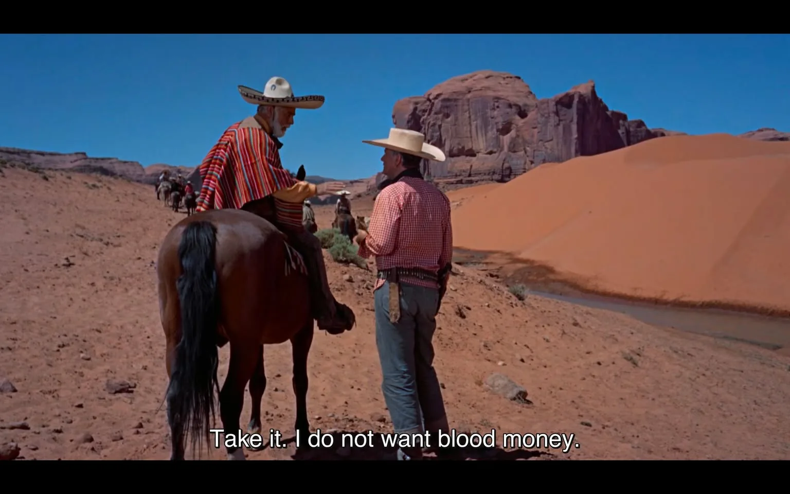 In a desert landscape, two men are engaged in a conversation. One man is seated on a brown horse, wearing a colorful striped poncho and a large sombrero, while the other stands beside him, dressed in a checkered shirt and a wide-brimmed hat. The background features striking red rock formations and sandy dunes under a clear blue sky. The seated man gestures with his hand, indicating a point of discussion, while the standing man appears to be holding something in his hands. The dialogue suggests a serious tone, with the seated man stating, "Take it. I do not want blood money." The setting is arid and expansive, emphasizing the isolation of the characters.