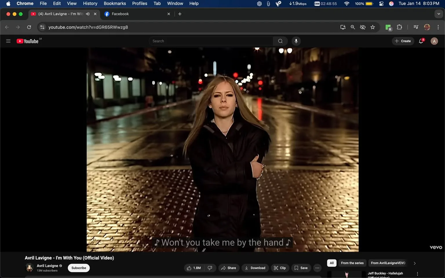 In the image, a person stands confidently in the middle of a wet, cobblestone street, illuminated by streetlights reflecting off the ground. The setting appears to be urban and nighttime, with a few buildings visible in the background. The individual is wearing a dark jacket and has long hair that flows with the wind. Their arms are crossed, and they have a serious expression on their face. The atmosphere is moody, enhanced by the wet pavement and dim lighting. The text overlay includes lyrics, indicating a connection to a song.