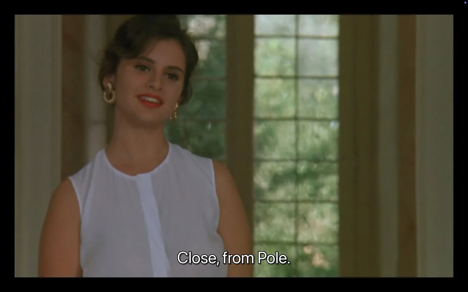 In the image, an unidentified woman stands smiling, wearing a white sleeveless top. Her hair is styled in soft waves, and she has large hoop earrings. The background features large windows that allow natural light to filter in, creating a bright atmosphere. The text at the bottom reads, "Close, from Pole," suggesting a dialogue or context related to her introduction. The setting appears to be indoors, likely in a spacious room with a relaxed ambiance. There are no visible brands or logos in the image.