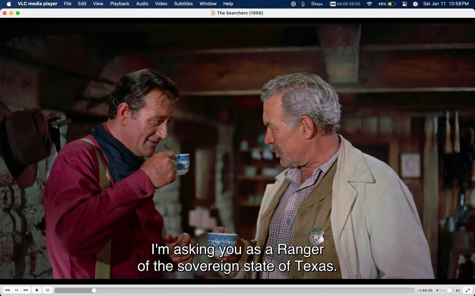 In the scene, two men are conversing in a rustic indoor setting, likely a cabin or similar structure. One man, dressed in a red shirt with a black scarf, is holding a small blue cup, appearing to take a sip while speaking. The other man, wearing a light-colored coat over a checkered shirt, listens intently. The dialogue suggests a serious discussion, with the first man stating, "I'm asking you as a Ranger of the sovereign state of Texas." The background features wooden beams and shelves, contributing to the cozy yet rugged atmosphere. There are no visible brands or logos in the scene.