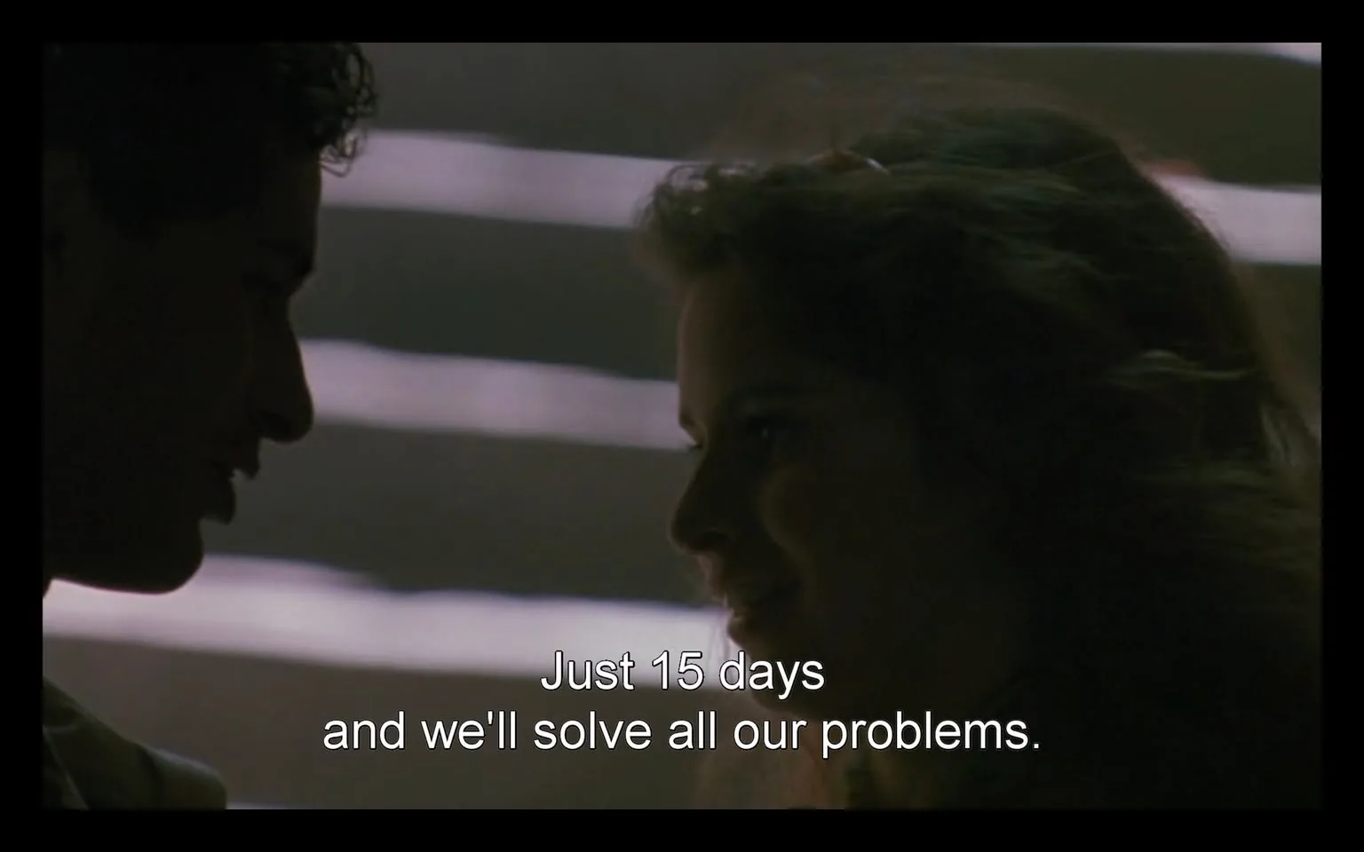In the image, two individuals are engaged in a close conversation, with one person appearing to speak directly to the other. The lighting is dim, creating a somewhat intimate atmosphere. The person on the left has dark, curly hair and is partially silhouetted, while the person on the right has lighter, wavy hair and is smiling. The text overlay reads, "Just 15 days and we'll solve all our problems," indicating a sense of urgency or hope in their dialogue. The background features soft, blurred lines, suggesting a setting that may be indoors, possibly with a focus on the two individuals rather than the environment.