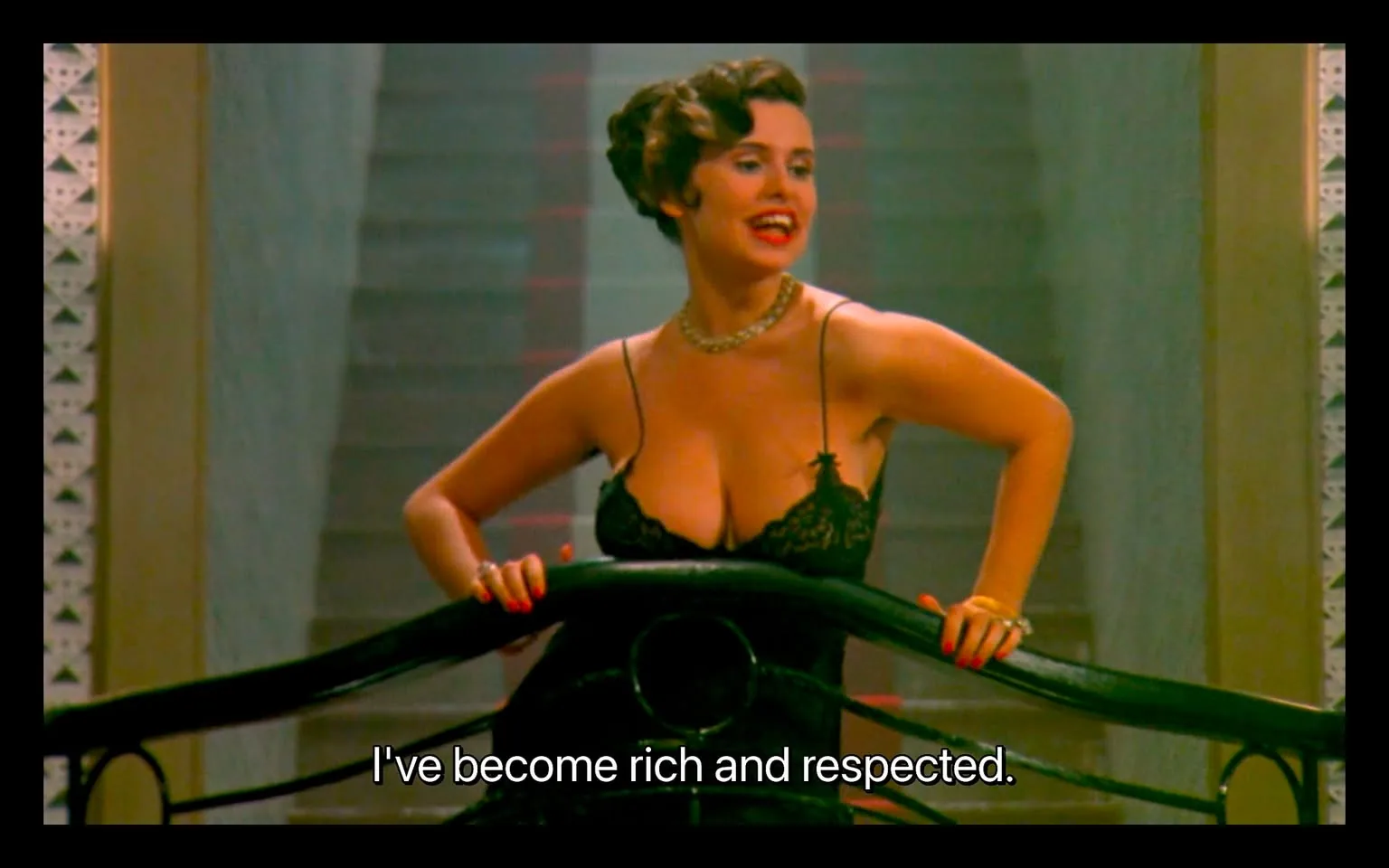 In the scene, an unidentified adult woman stands confidently on a staircase, leaning against a railing. She wears a black lace outfit that is revealing, accentuating her figure. Her hair is styled in vintage waves, and she has a bright smile, suggesting a lively demeanor. The background features a staircase with a soft focus, indicating an indoor setting. The woman is speaking, as indicated by the text overlay that reads, "I've become rich and respected." The overall atmosphere appears theatrical, possibly from a film or performance, with a focus on her expression and posture.