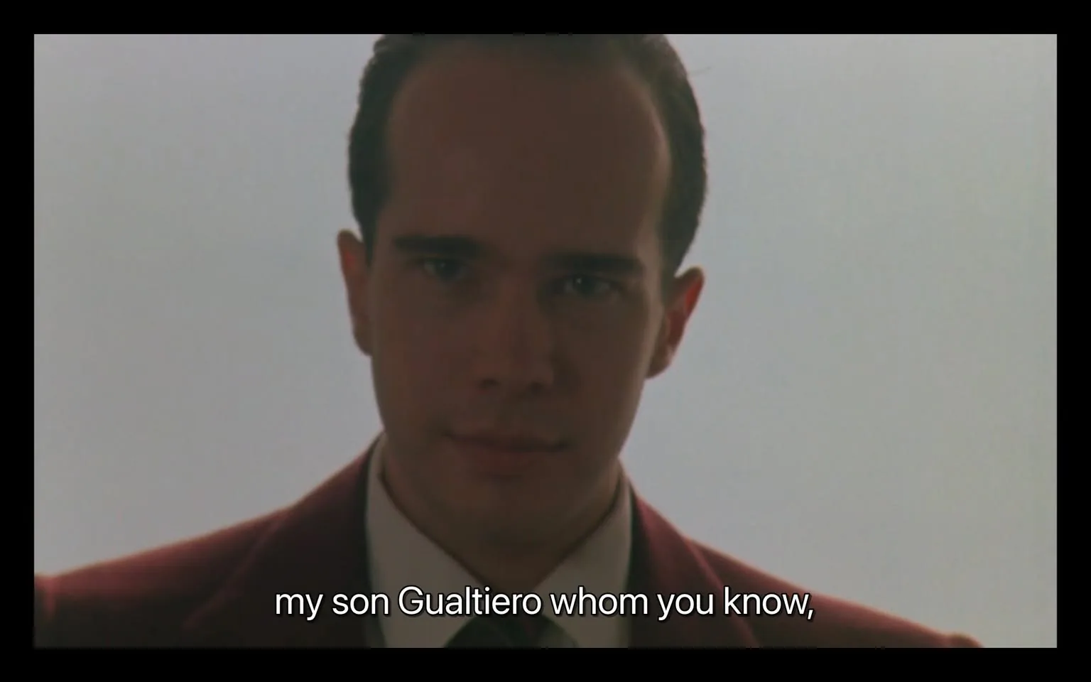 In the image, a young man with a slight smile is looking directly at the camera. He has short, neatly styled hair and is dressed in a dark suit with a white shirt and a tie. The background is a soft, light color, creating a minimalistic atmosphere. Below him, there is readable text that states, "my son Gualtiero whom you know," indicating a personal connection or introduction. The overall tone of the image is formal, suggesting a serious or significant moment. There are no visible brands, logos, or any elements indicating violence or sexual content.