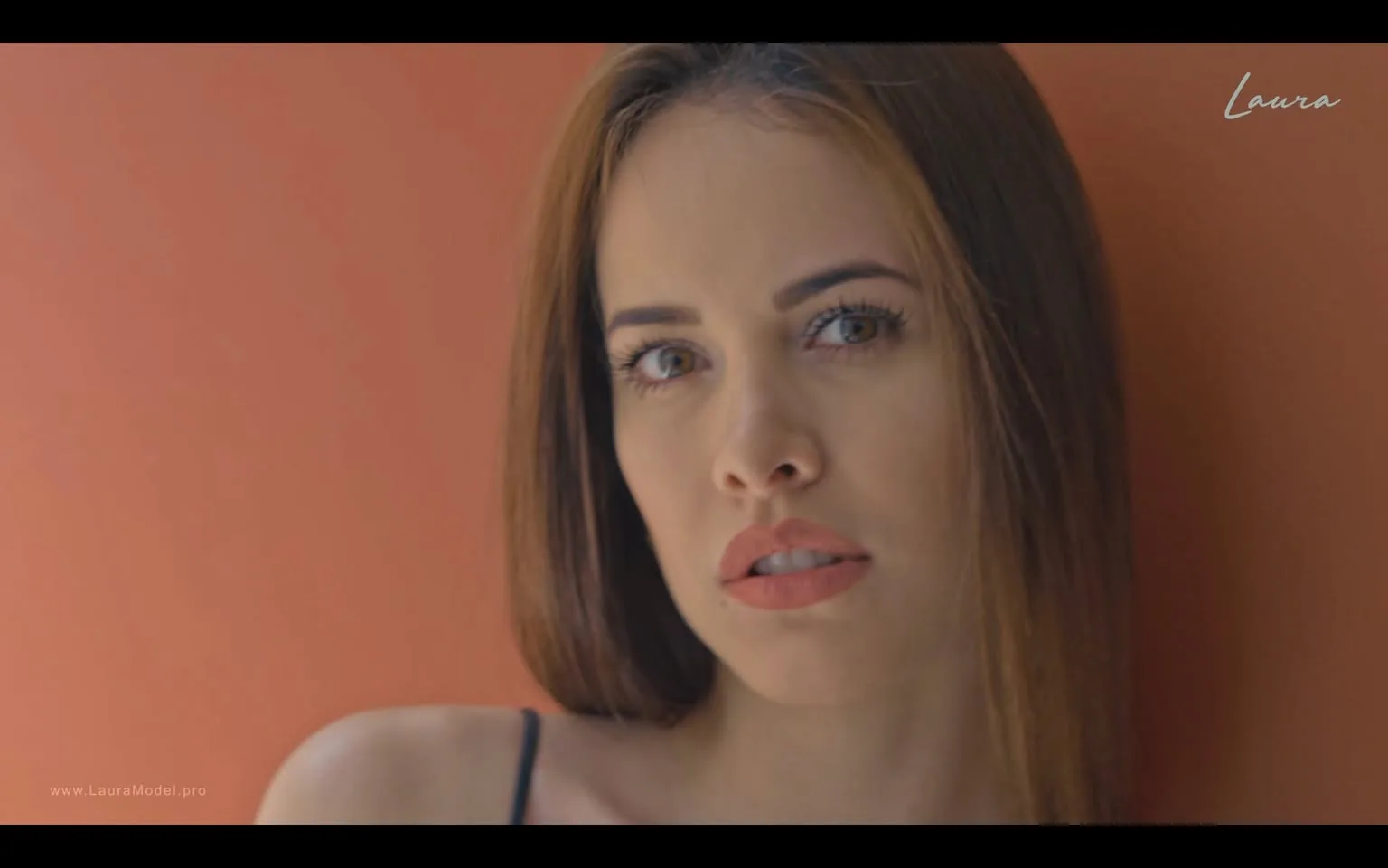 In the image, a woman with long, straight hair is positioned against a warm, orange background. She has a neutral expression, with her gaze directed slightly off-camera. Her makeup is subtle, highlighting her features, including her full lips and defined eyebrows. She is wearing a black spaghetti strap top, which contrasts with the background. The lighting is soft, creating a gentle ambiance that enhances her facial features. There are no visible logos or text in the image, and the overall composition focuses on her expression and the color contrast with the background.