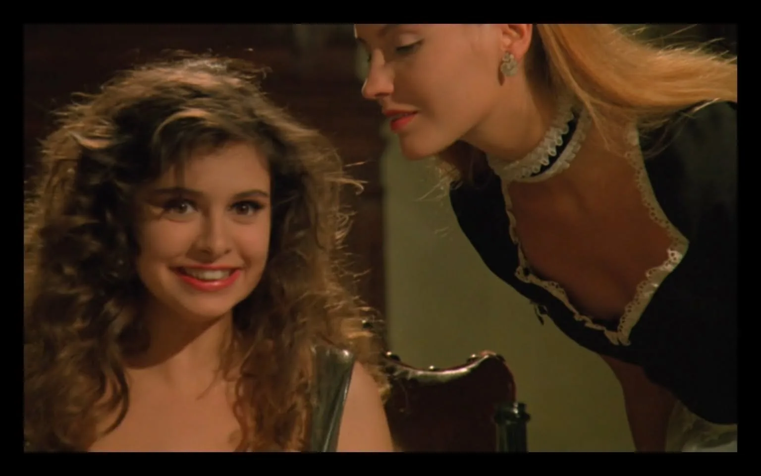 In the scene, two women are engaged in a close conversation. The first woman, with curly hair and a bright smile, appears to be in a casual setting, possibly at a dining table. She is wearing a dark outfit that complements her youthful appearance. The second woman, leaning in closely, has long, straight hair and is dressed in a black outfit with white lace accents, suggesting a role of service or hospitality. The atmosphere seems warm and friendly, with the first woman displaying a cheerful expression. The background is softly lit, indicating an indoor setting, likely during the evening. There are no visible brands or logos in the image. The interaction between the two women hints at a personal or light-hearted exchange.