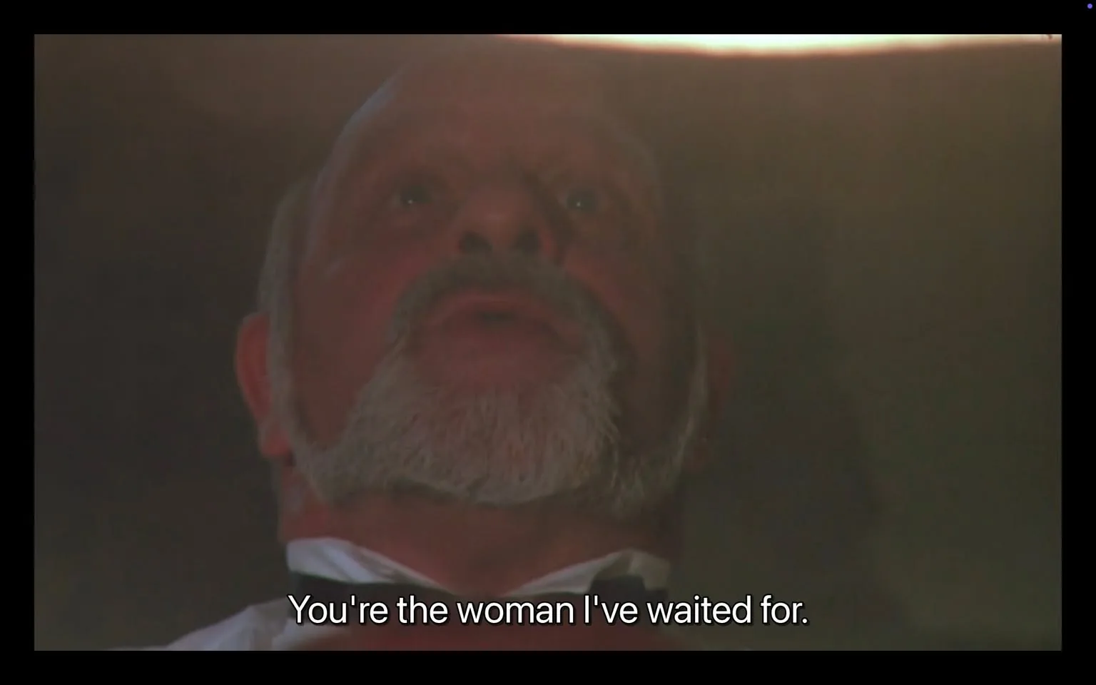 In the image, an unidentified adult male with a prominent beard and a serious expression is speaking. His face is illuminated from above, creating a dramatic effect. He appears to be in a tense or emotional moment, as indicated by his dialogue: "You're the woman I've waited for." The background is dimly lit, which adds to the intensity of the scene. There are no visible brands or logos present, and the setting does not suggest any overt violence or sexual content. The overall atmosphere hints at a significant narrative moment, possibly from a film or theatrical performance.