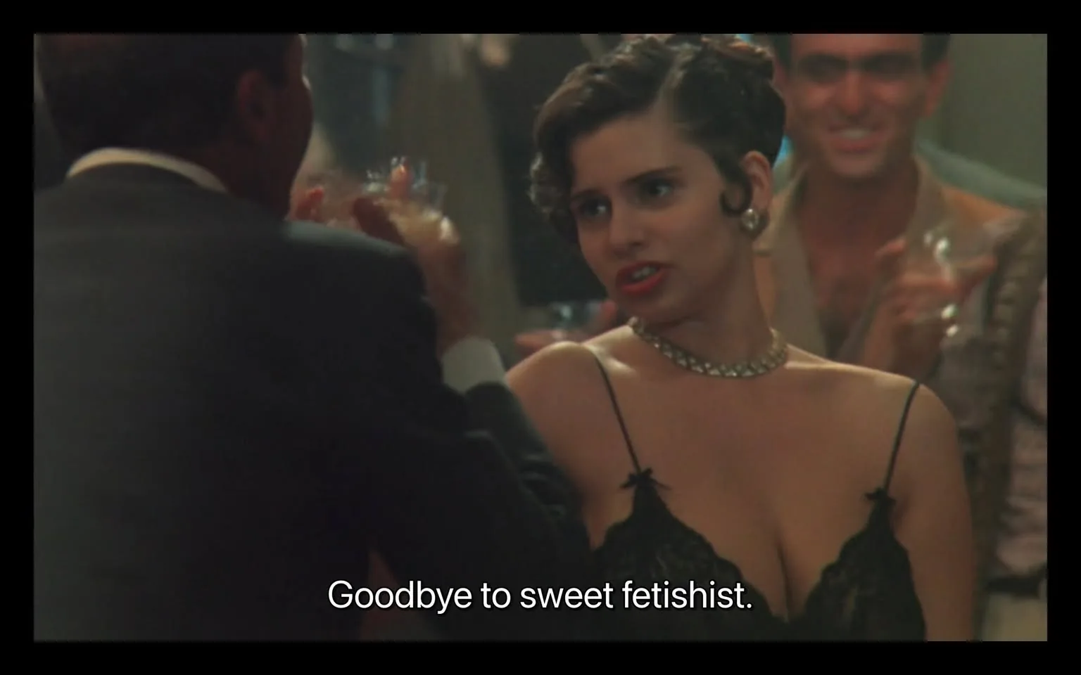 In a lively social setting, an unidentified adult woman engages in conversation with another individual, who is partially obscured. The woman has a vintage hairstyle, styled in soft curls, and wears a black lace dress with thin straps, which accentuates her figure. She appears animated, with a confident expression, as she says, "Goodbye to sweet fetishist." The background features other individuals, some of whom are smiling and holding drinks, suggesting a festive atmosphere. The lighting is warm, typical of a bar or club environment, enhancing the intimate vibe of the scene. The presence of drinks indicates a celebratory mood, but the context of the conversation hints at a more complex interaction.