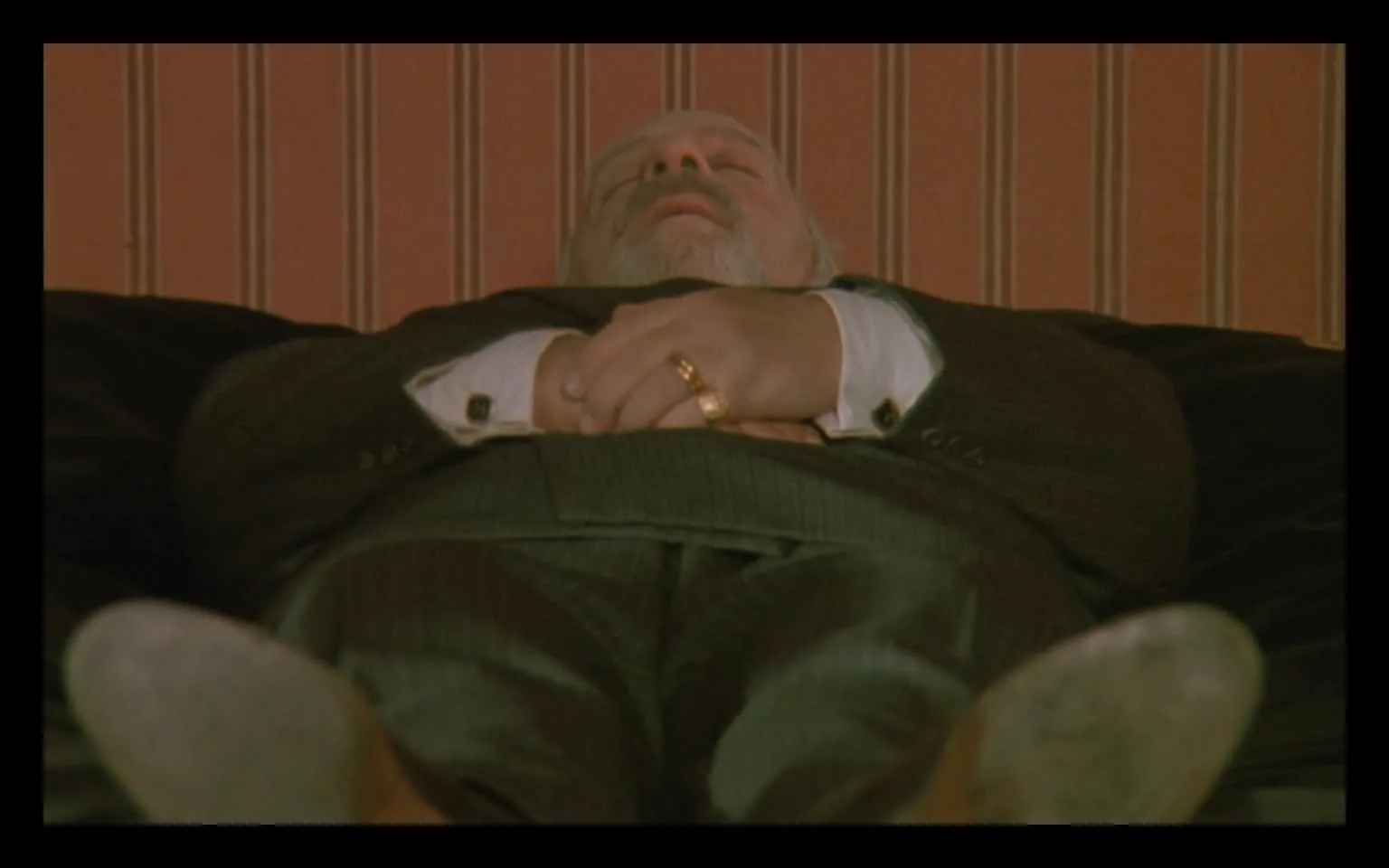 In the image, an unidentified adult male is lying on a couch, appearing to be in a state of rest. He is dressed in a dark suit with a white shirt, and his hands are clasped together over his abdomen. A gold ring is visible on one of his fingers. The background features a striped wall in shades of pink and beige, contributing to an indoor setting. The man's head is tilted back slightly, and his eyes are closed, suggesting he may be asleep or resting. The overall atmosphere conveys a sense of calm or inactivity.