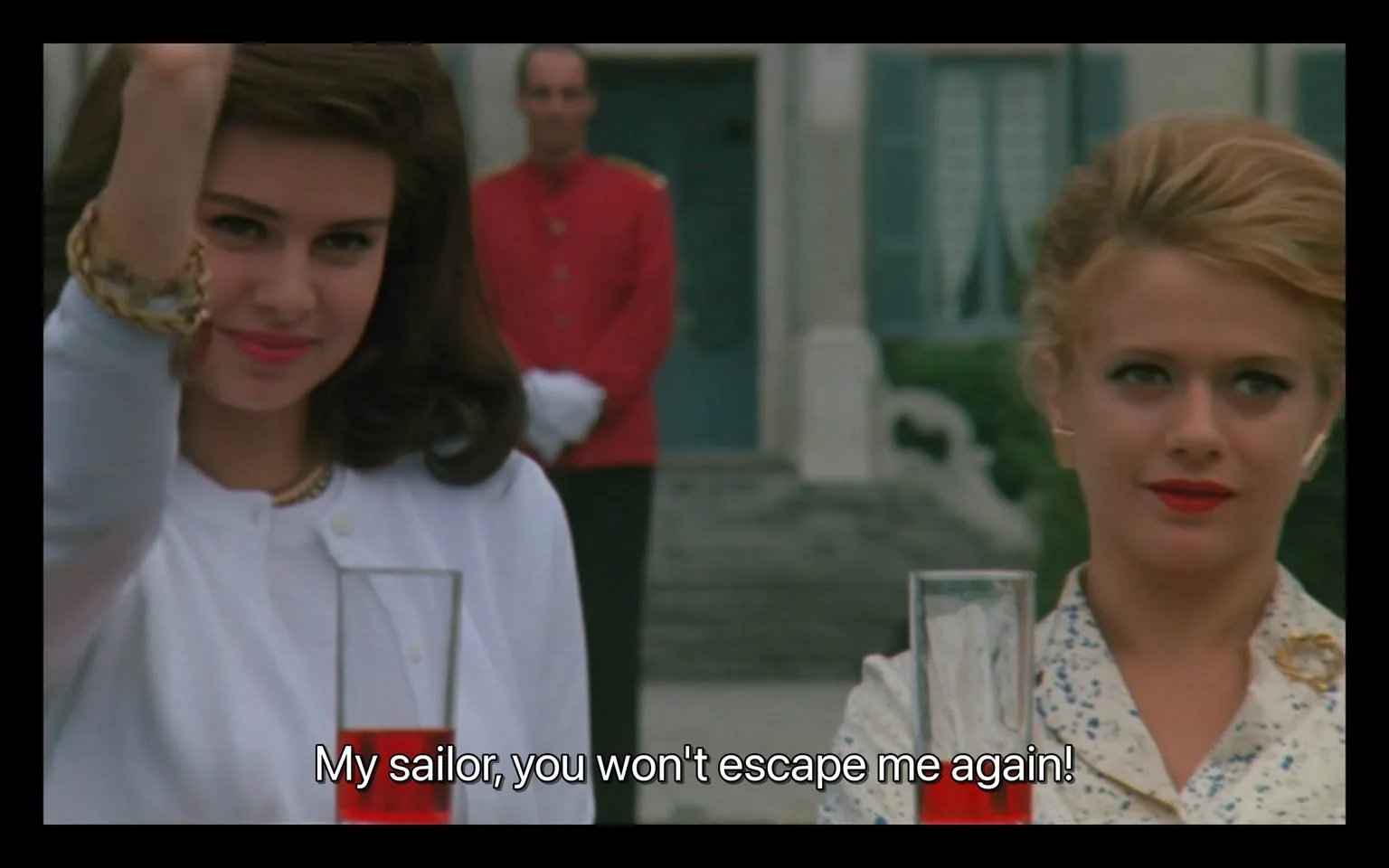 In the image, two women are prominently featured in the foreground, each holding a tall glass filled with a red beverage. The woman on the left has dark, voluminous hair and is wearing a white top, adorned with a gold bracelet. She appears to be playfully raising her hand, possibly in a gesture of greeting or emphasis. The woman on the right has lighter hair styled in an elegant updo and is dressed in a patterned outfit, with a confident expression. Behind them, a man in a red uniform stands, seemingly observing the scene. The setting appears to be outdoors, with a building and windows visible in the background, suggesting a formal or social event. The text overlay reads, "My sailor, you won't escape me again!" indicating a playful or teasing interaction between the women and someone not visible in the image.