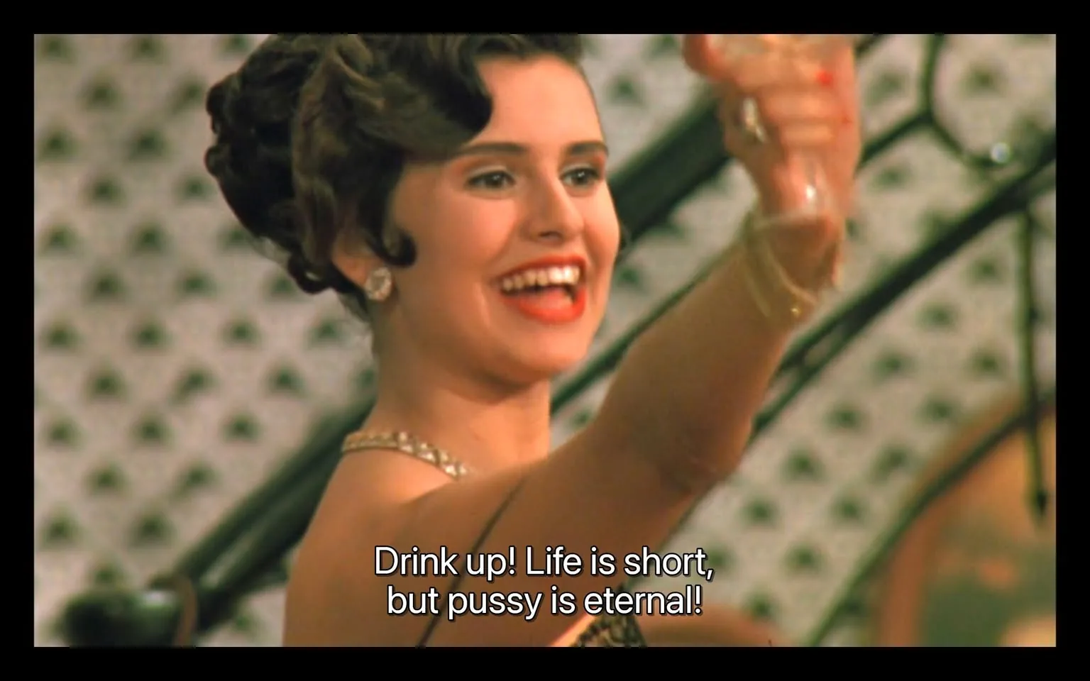 In a lively setting, an unidentified woman is seen joyfully raising a glass, her expression bright and animated. She has dark, styled hair and is wearing a glamorous outfit adorned with jewelry, including earrings and a necklace. The background features a patterned wall, suggesting an indoor event, likely a party or celebration. The text overlay reads, "Drink up! Life is short, but pussy is eternal!" indicating a playful and bold atmosphere. The scene captures a moment of revelry, with the woman exuding confidence and enthusiasm. There are no visible brands or logos in the image.