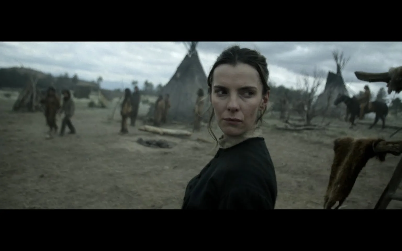 In the foreground, an unidentified woman stands with a serious expression, glancing back over her shoulder. She has long, dark hair and is dressed in a dark, simple garment that appears to be from a historical period. The background features several figures, likely in traditional attire, moving about a settlement with teepees visible. The environment is outdoors, characterized by a barren landscape under a cloudy sky, suggesting a somber atmosphere. The overall scene conveys a sense of tension or unease, with the woman’s expression hinting at concern or vigilance.