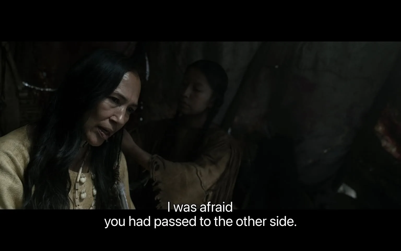 In the scene, two individuals are present in a dimly lit setting that appears to be indoors, possibly in a tent or a cave. One woman, with long dark hair and wearing a light-colored garment, is speaking with a concerned expression. She seems to be addressing another person, who is partially visible in the background. This second individual, also a woman, has her hair tied back and is dressed in a similar style, suggesting a cultural or historical context. The dialogue indicates a sense of worry, as the first woman expresses her fear that the other has "passed to the other side," implying a serious or life-threatening situation. The overall atmosphere conveys a deep emotional connection between the characters, set against a backdrop that hints at a challenging environment.