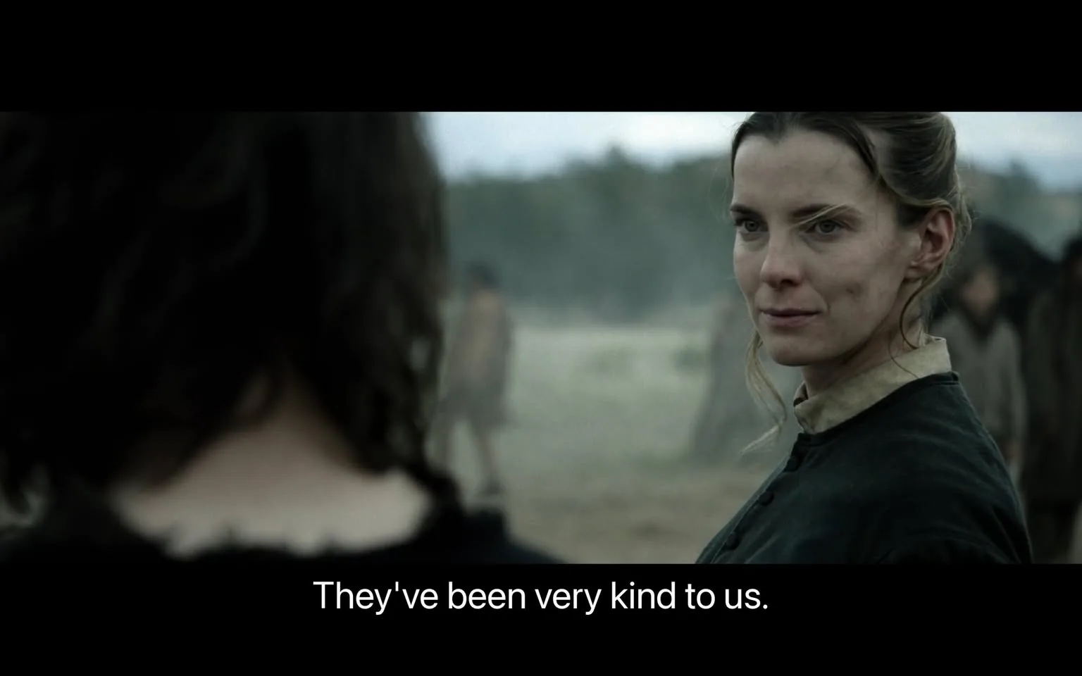 In the image, a woman with long, light brown hair is prominently featured, looking directly at another person off-screen. She has a serious expression, with slight dimples visible on her cheeks, suggesting a contemplative or reflective mood. The woman is dressed in a dark, simple garment, typical of a historical or period setting. The background is blurred, but hints at a desolate landscape with a few indistinct figures in the distance, possibly indicating a rural or wilderness environment. The dialogue displayed at the bottom reads, "They've been very kind to us," suggesting a conversation about kindness or hospitality. The overall atmosphere appears somber and reflective, aligning with the themes often found in dramatic narratives.