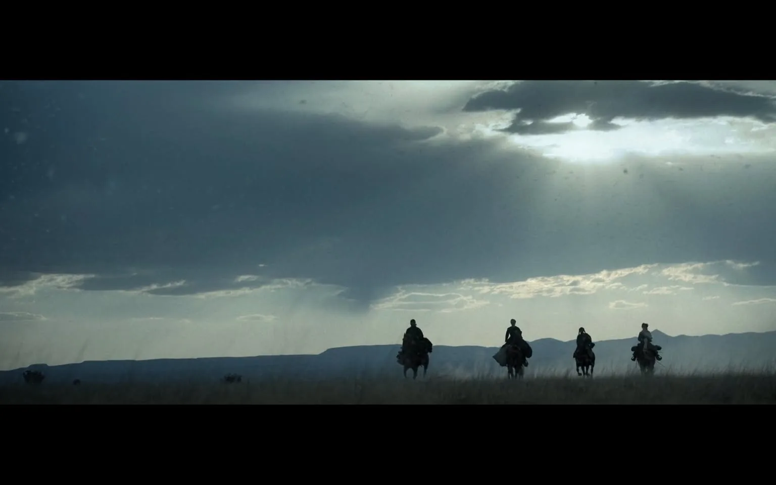 In the image, four riders on horseback traverse a vast, open landscape under a dramatic sky filled with clouds. The scene is set during what appears to be late afternoon or early evening, as the light casts a soft glow across the horizon. The riders are silhouetted against the backdrop of distant mountains, creating a striking contrast with the darker clouds above. The overall atmosphere is one of tranquility and adventure, evoking a sense of exploration in a natural setting. The grass in the foreground sways gently, hinting at a breeze, while the expansive sky adds depth to the composition.