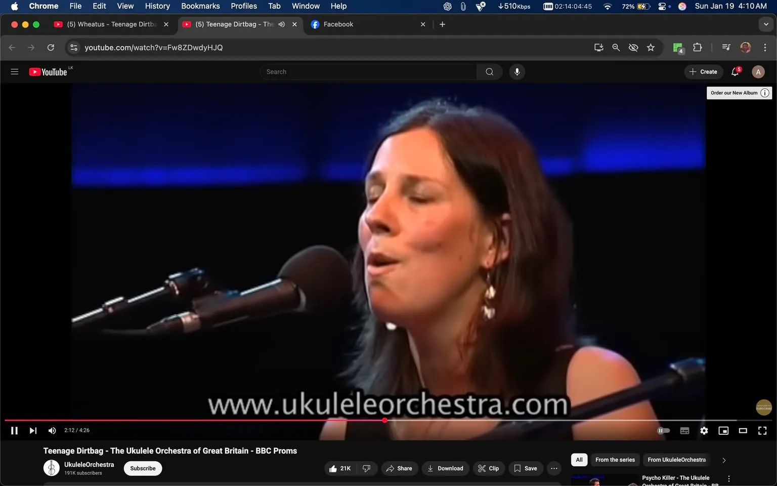 In the image, a woman is captured mid-performance, singing into a microphone. She has long, dark hair and is wearing a black top. Her expression is focused and emotive, with her eyes closed as she engages with the music. The background features a soft blue lighting that creates an atmospheric stage setting, suggesting this is a live performance, possibly at a concert or music festival. The microphone is positioned close to her mouth, indicating she is actively singing. There are no visible logos or brands in the image, and the overall mood appears to be one of artistic expression. The context suggests this is part of a performance by the Ukulele Orchestra of Great Britain, as indicated by the text overlay on the video.