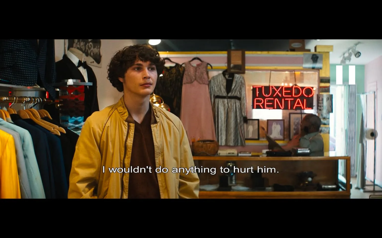 In the scene, a young man with curly hair stands in a clothing rental shop, wearing a yellow jacket over a brown shirt. He appears contemplative, with a serious expression as he speaks the line, "I wouldn't do anything to hurt him." The shop is filled with various garments, including tuxedos and dresses, with a prominent neon sign reading "TUXEDO RENTAL" in the background. The setting is indoors, likely during the day, as indicated by the bright lighting. 

Behind the young man, several suits are displayed on mannequins, and there are racks of clothing, including a mix of colors like blue and yellow. A person can be seen in the background, possibly an employee, engaged in a phone conversation. The overall atmosphere suggests a moment of personal conflict or concern, highlighted by the young man's earnest demeanor. There are no visible signs of violence, sexual content, or any other sensitive material in this scene.