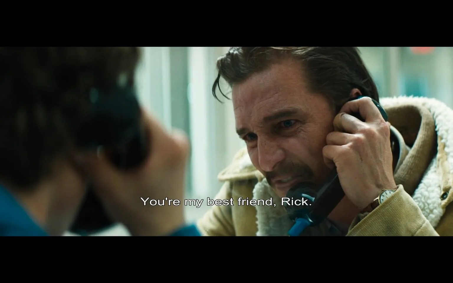 In the scene, a man with a serious expression is speaking on the phone, conveying a strong emotional message. He appears to be in a tense situation, emphasizing the words, "You're my best friend, Rick." His face shows signs of distress, with furrowed brows and a focused gaze. He is dressed in a light-colored jacket with a hood, suggesting a casual yet somewhat rugged appearance. The background is slightly blurred, indicating that the focus is on the man and the phone call. 

The other person on the line is not fully visible, but their hand is seen holding a black phone receiver, suggesting they are engaged in the conversation. The setting appears to be indoors, possibly in a public place like a phone booth or a waiting area, with soft lighting that adds to the emotional tone of the moment. The dialogue hints at a deep connection between the two characters, suggesting themes of loyalty and friendship amidst a challenging situation.