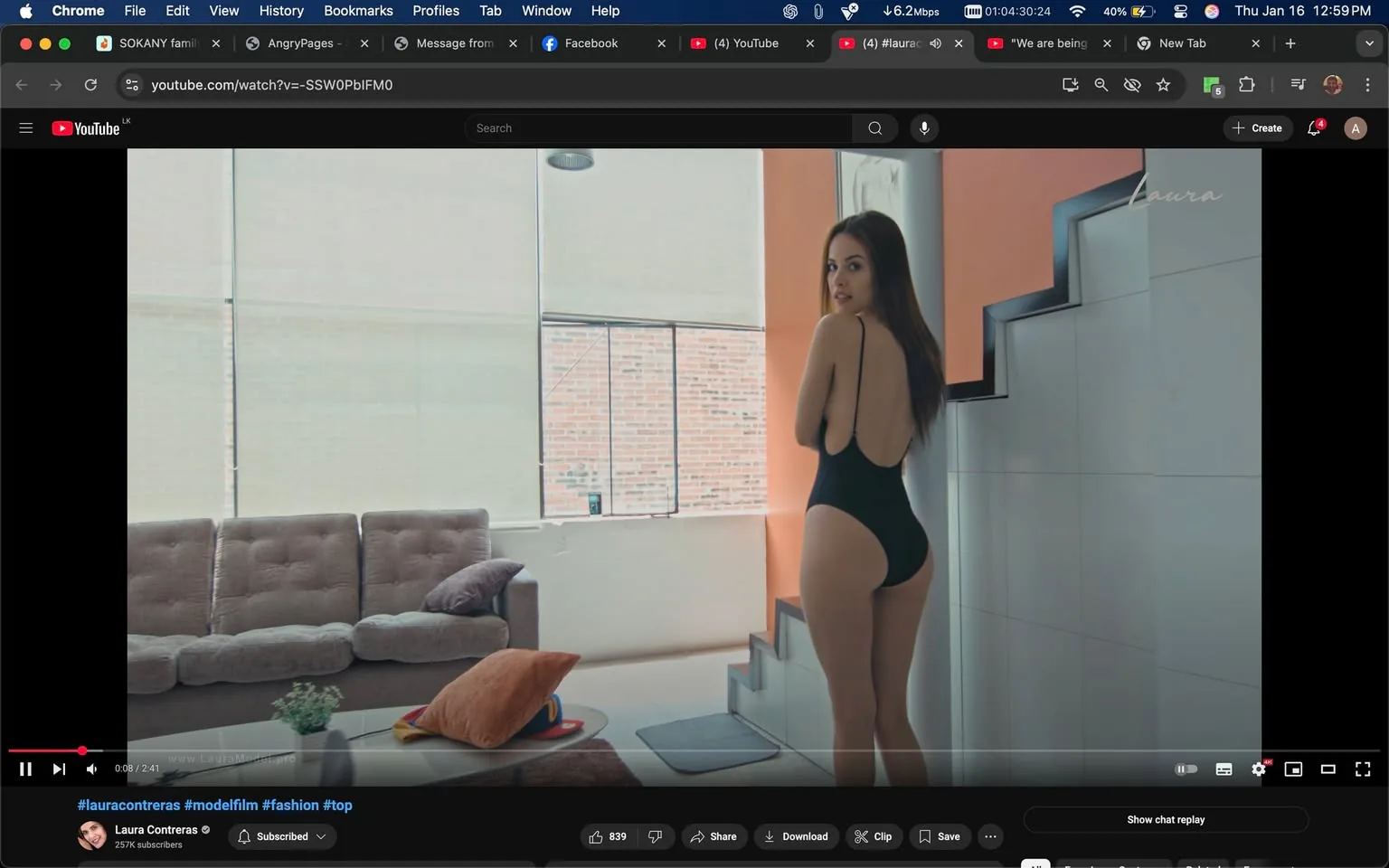 In the image, an unidentified adult is posing in a stylish indoor setting, showcasing a modern aesthetic. The individual is wearing a black swimsuit with a low back, standing near a staircase. The background features large windows allowing natural light to flood the space, highlighting a cozy living area with a gray sofa and colorful cushions. A small plant is placed on a table, adding a touch of greenery to the scene. The overall ambiance is relaxed and contemporary, suggesting a fashion or modeling context.