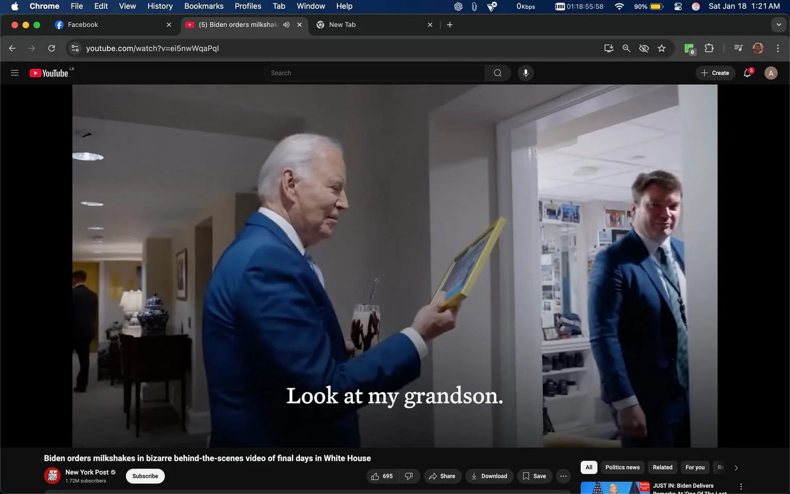 In the image, an older adult is seen holding a framed picture, smiling as he shows it to another individual in a suit standing in a doorway. The older adult is dressed in a blue suit and has white hair, while the other person appears to be in a dark suit with a light-colored tie. The setting is indoors, likely within an office or a home, with soft lighting and a few decorative items visible in the background, including a lamp and a table. A glass with a drink is also held by the older adult. The text overlay reads, "Look at my grandson," suggesting a personal moment being shared.