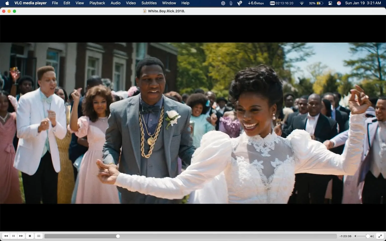A vibrant outdoor wedding scene unfolds, filled with joy and celebration. In the foreground, an unidentified adult woman in a stunning white gown with intricate lace details beams with happiness as she dances, holding hands with an unidentified adult man dressed in a gray suit adorned with gold chains. The crowd behind them, consisting of various individuals in colorful attire, also participates in the festivities, with some dancing and others smiling. The setting is bright and sunny, suggesting a cheerful atmosphere typical of a wedding. The background features a building and trees, enhancing the outdoor ambiance of the event.