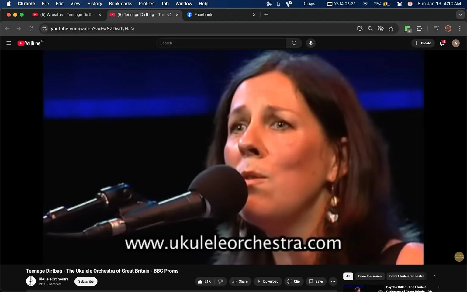 In the image, an unidentified adult woman is singing into a microphone, displaying a focused expression. She has long, dark hair and is wearing earrings. The setting appears to be a stage, likely during a performance, with a blue background that suggests a spotlight effect. The microphone is positioned close to her mouth, indicating she is actively engaged in singing. Below her, there is visible text promoting the website "www.ukuleleurchestra.com," which suggests she may be part of a musical group. The overall atmosphere is one of a live music performance, with an audience likely present, although they are not visible in the image.