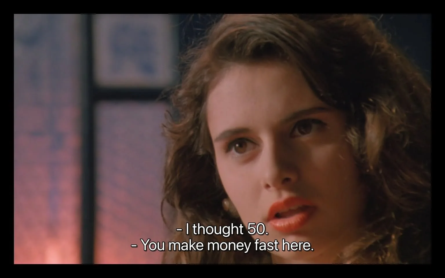 In the image, a woman with long, wavy hair and a serious expression is engaged in a conversation. She has prominent features, including full lips painted in a bold shade of red, and her eyes convey intensity. The background appears to be dimly lit, with hints of a colorful setting that suggests an indoor environment. The dialogue displayed on the screen indicates a discussion about money, with phrases like "I thought 50" and "You make money fast here," suggesting a transactional context. The overall mood seems tense, reflecting the seriousness of the conversation.