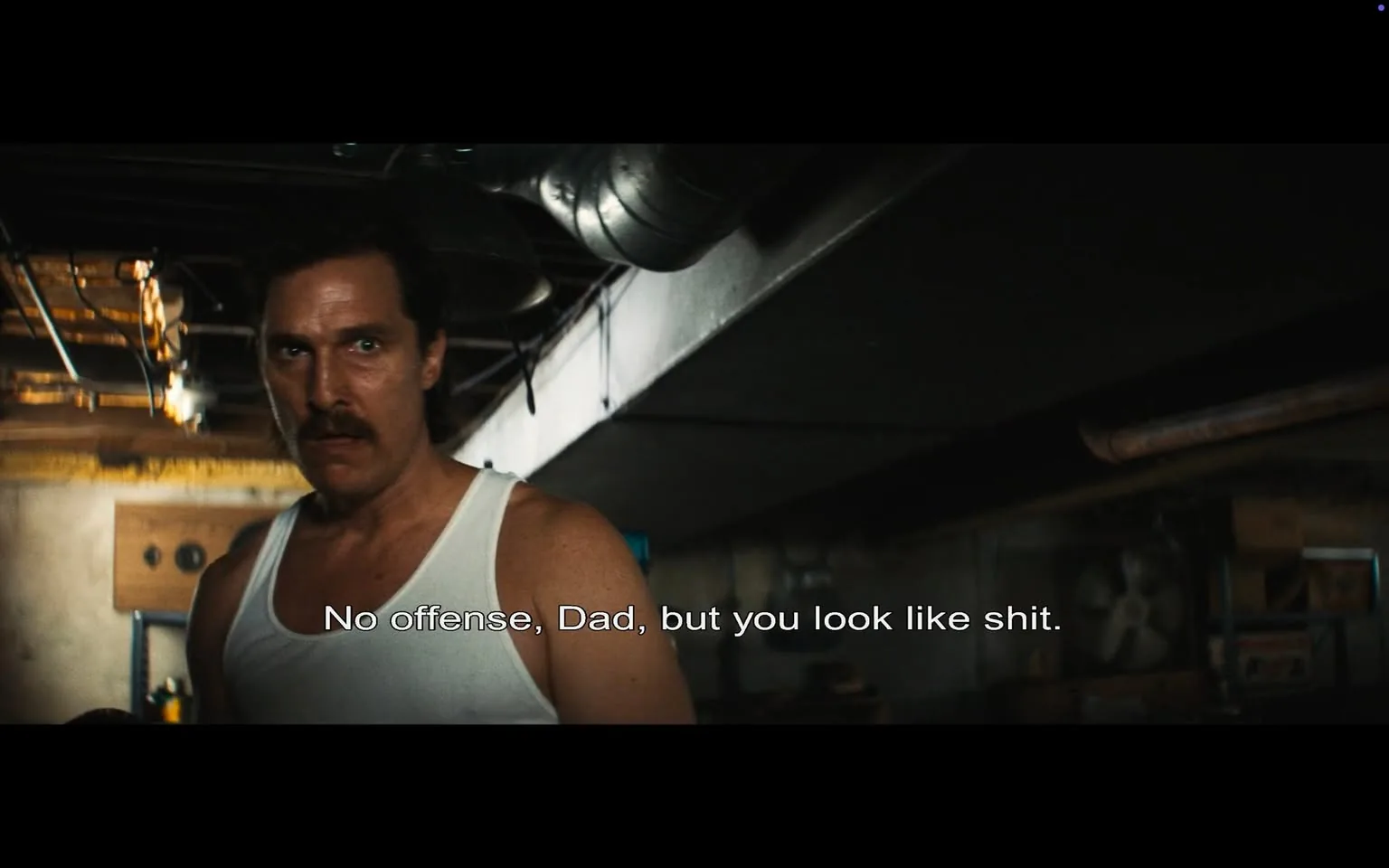 In a dimly lit basement, an unidentified adult male stands with a serious expression, wearing a white tank top. His hair is dark and slightly tousled, and he has a prominent mustache. The background reveals a cluttered space with various items, including boxes and a fan, suggesting a utilitarian environment. The dialogue displayed on the screen reads, "No offense, Dad, but you look like shit," indicating a personal and possibly confrontational moment between him and another character. The overall atmosphere feels tense, with the lighting casting shadows that enhance the seriousness of the scene.