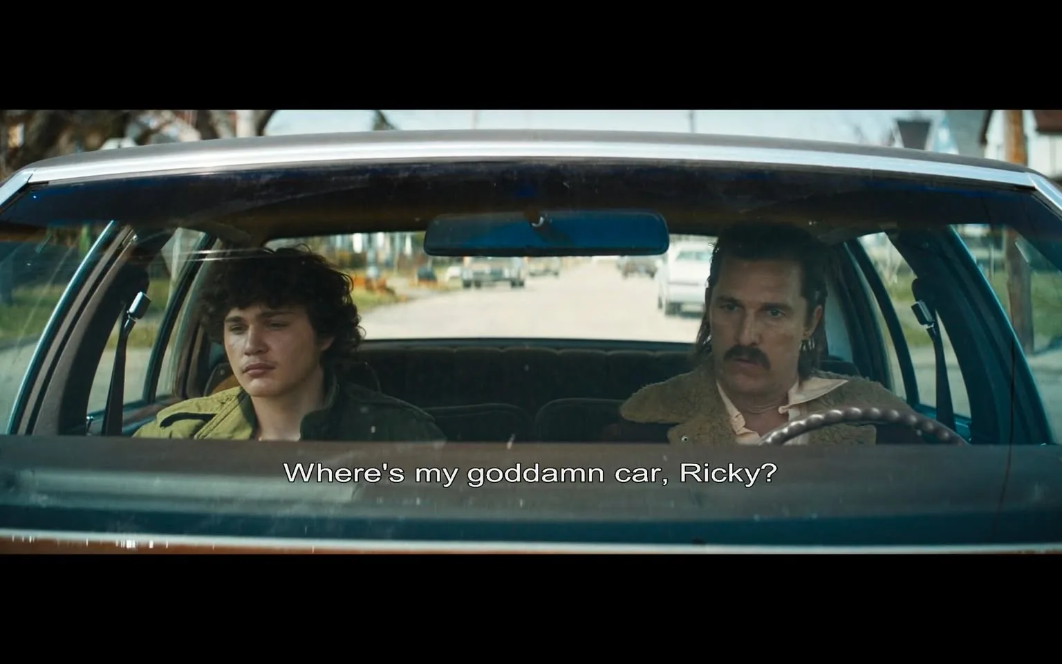 In the car, two characters are seated in the front seats, with the driver on the right and a passenger on the left. The driver has a mustache and is wearing a light-colored shirt with a fur-lined jacket, conveying a rugged appearance. He looks serious and is directing a frustrated question at the passenger, asking, "Where's my goddamn car, Ricky?" The passenger, who appears younger with curly hair, looks contemplative and somewhat troubled, not making eye contact with the driver. 

The setting is outdoors, as indicated by the view through the windshield, which shows a street lined with trees and parked cars. The atmosphere seems tense, suggesting a moment of conflict or frustration between the two characters. The car itself has a vintage look, with a classic dashboard and interior design typical of older models. There are no visible brands or logos in the image, and the focus remains on the characters and their interaction.