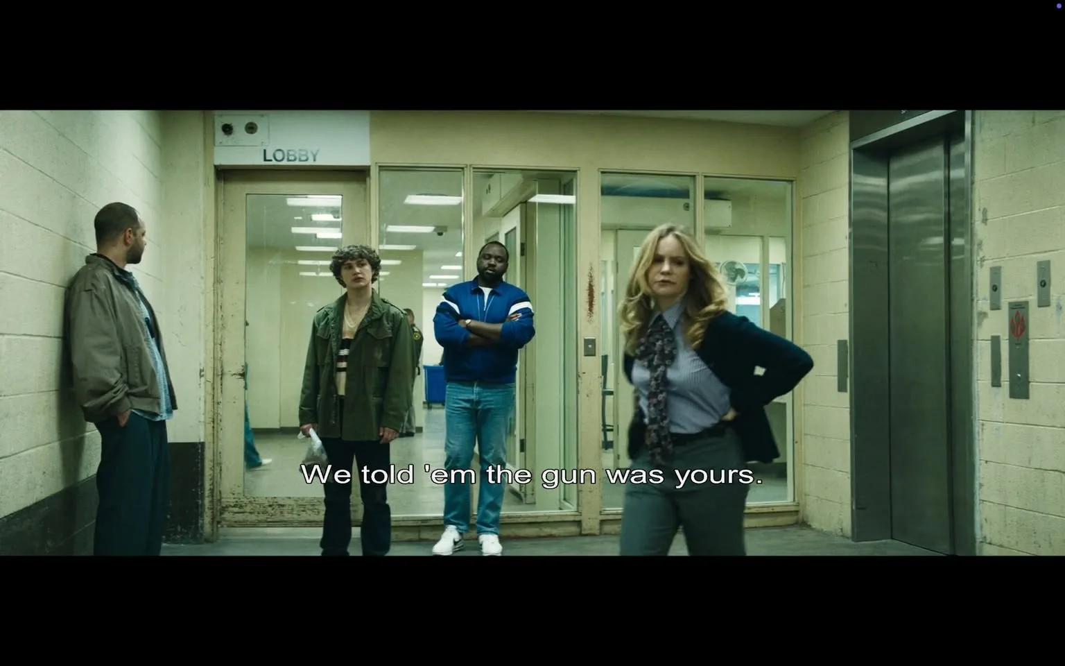In this scene, four characters are present in a stark, indoor setting that resembles a police station or a similar facility. The atmosphere is tense, indicated by the dialogue displayed on the screen: "We told 'em the gun was yours." The characters include a man leaning against the wall on the left, who appears to be in his 30s, dressed in a casual outfit with a gray jacket. Next to him stands a younger individual, possibly a teenager, wearing a green jacket over a striped shirt, looking concerned. In the center, a man in a blue jacket with white stripes stands with his arms crossed, exuding a sense of authority or defiance. To the right, a woman in a blue blouse and dark pants, with long hair, appears assertive as she places her hands on her hips.

The setting features a glass door labeled "LOBBY," with visible fluorescent lighting overhead, suggesting a daytime scenario. The walls are plain and somewhat worn, contributing to the serious tone of the scene. There are no visible brands or logos, but the presence of a gun is implied through the dialogue, indicating a potential conflict or accusation among the characters. The expressions and body language of the characters suggest a high-stakes conversation, possibly involving legal or criminal implications.