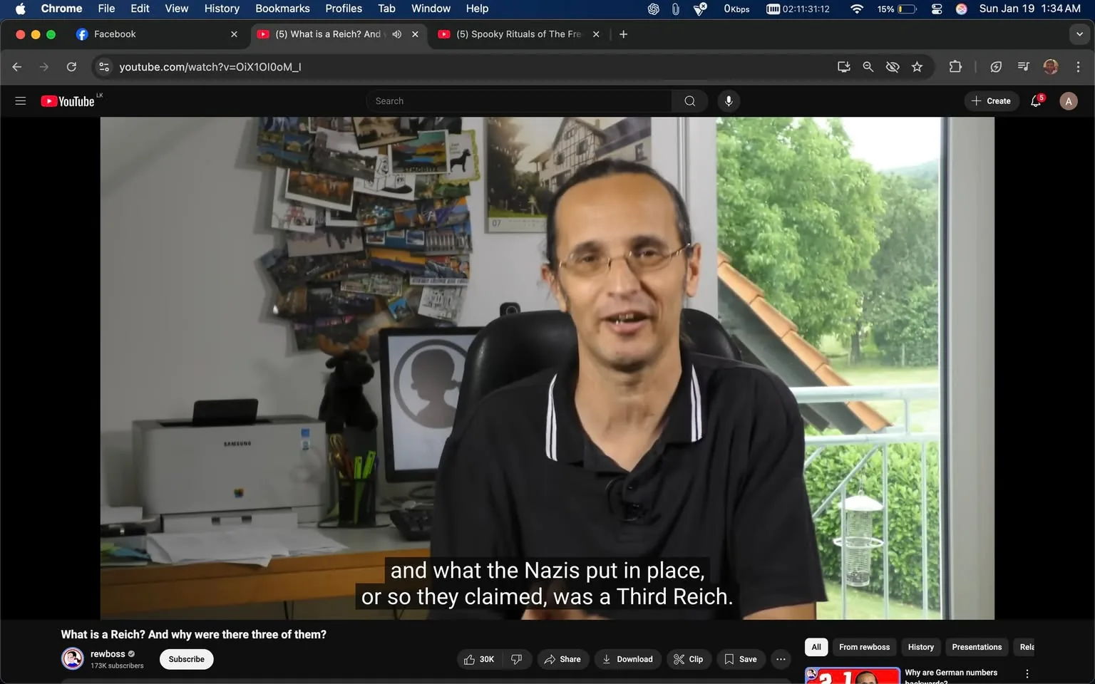 In the video, a man is seated in an office-like setting, speaking directly to the camera. He has a slight smile and appears engaged in his discussion about historical topics, specifically mentioning the Nazis and the concept of a Third Reich. He is wearing a black polo shirt with white stripes on the collar. Behind him, a wall is adorned with various postcards and photographs, suggesting a personal touch to the workspace. 

To his left, a printer is visible, branded with the Samsung logo, and there are several papers scattered on the desk, indicating an active work environment. The background features a window with a view of greenery outside, suggesting it is daytime. The overall atmosphere is casual and informative, with no signs of violence, sexual content, or any political symbols. The video appears to be part of a YouTube channel named "rewboss," which has a subscriber count of 173,000.