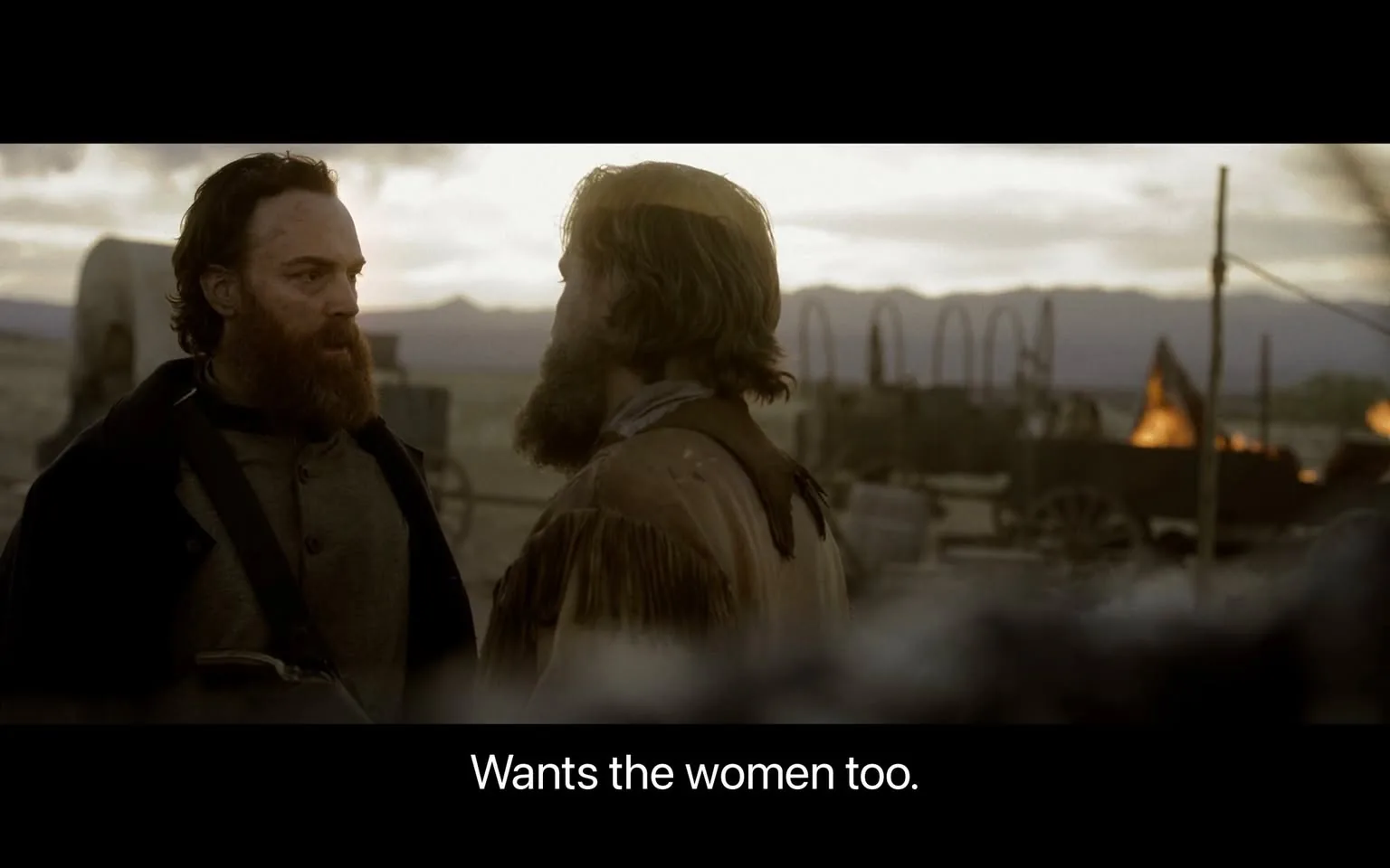 In the scene, two men are engaged in a serious conversation, likely discussing a contentious issue. One man has a long, dark beard and is dressed in a dark coat, while the other has a lighter beard and is wearing a fringed jacket, indicative of a rugged, possibly historical setting. The background features a desolate landscape with mountains in the distance and some wagons, suggesting a frontier or western theme. The lighting is soft, hinting at either dawn or dusk, which adds to the dramatic atmosphere. The dialogue displayed at the bottom of the image indicates a desire for control or possession, reflecting themes of conflict and power dynamics.