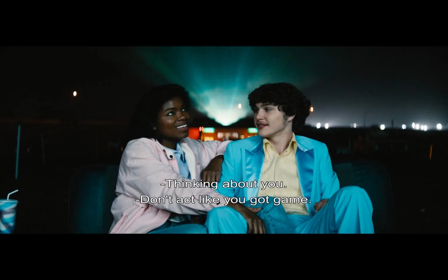 In the scene, two young characters are seated together on a couch, engaging in a playful conversation. The female character, wearing a light pink jacket over a blue top, has a bright smile and appears to be enjoying the moment. She leans slightly towards the male character, who is dressed in a light blue suit with a yellow shirt underneath. His expression is relaxed, and he seems to be responding to her with a hint of amusement. 

The background is dimly lit, suggesting it might be nighttime, with soft beams of light illuminating the area behind them, possibly from a nearby event or venue. There are hints of a festive atmosphere, indicated by the colorful lights in the distance. The dialogue displayed on the screen suggests a flirtatious exchange, with the female character teasing the male about his confidence. 

No logos, brands, or sensitive content are visible in this scene. The overall mood is light and youthful, capturing a moment of connection between the two characters.