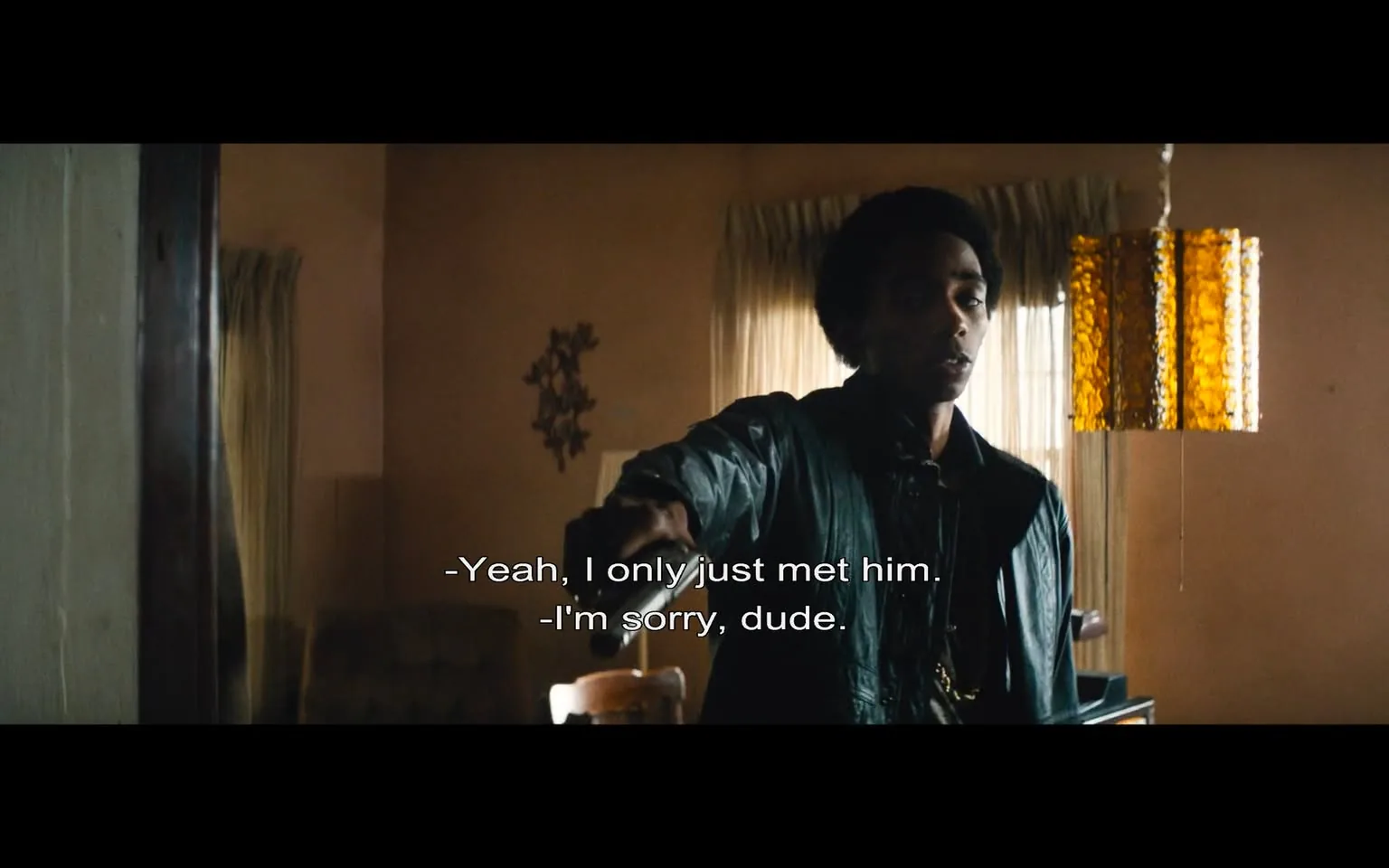 In this scene, a young man stands in a room, holding a gun, indicating a moment of tension or confrontation. He appears to be in his late teens or early twenties, wearing a dark jacket and a chain around his neck. His expression is serious, and he seems to be engaged in a conversation, possibly defending himself or explaining a situation. The dialogue suggests he has just met someone, and there is an apology from another person, indicating a sense of regret or concern.

The setting is an indoor space with a warm color palette, featuring light brown walls and a window with sheer curtains that allow some natural light to filter in. A distinctive yellow-orange pendant light hangs from the ceiling, adding to the room's ambiance. In the background, there is a wooden table and a chair, suggesting a casual living area. The overall atmosphere conveys a mix of urgency and unease, heightened by the presence of the weapon.