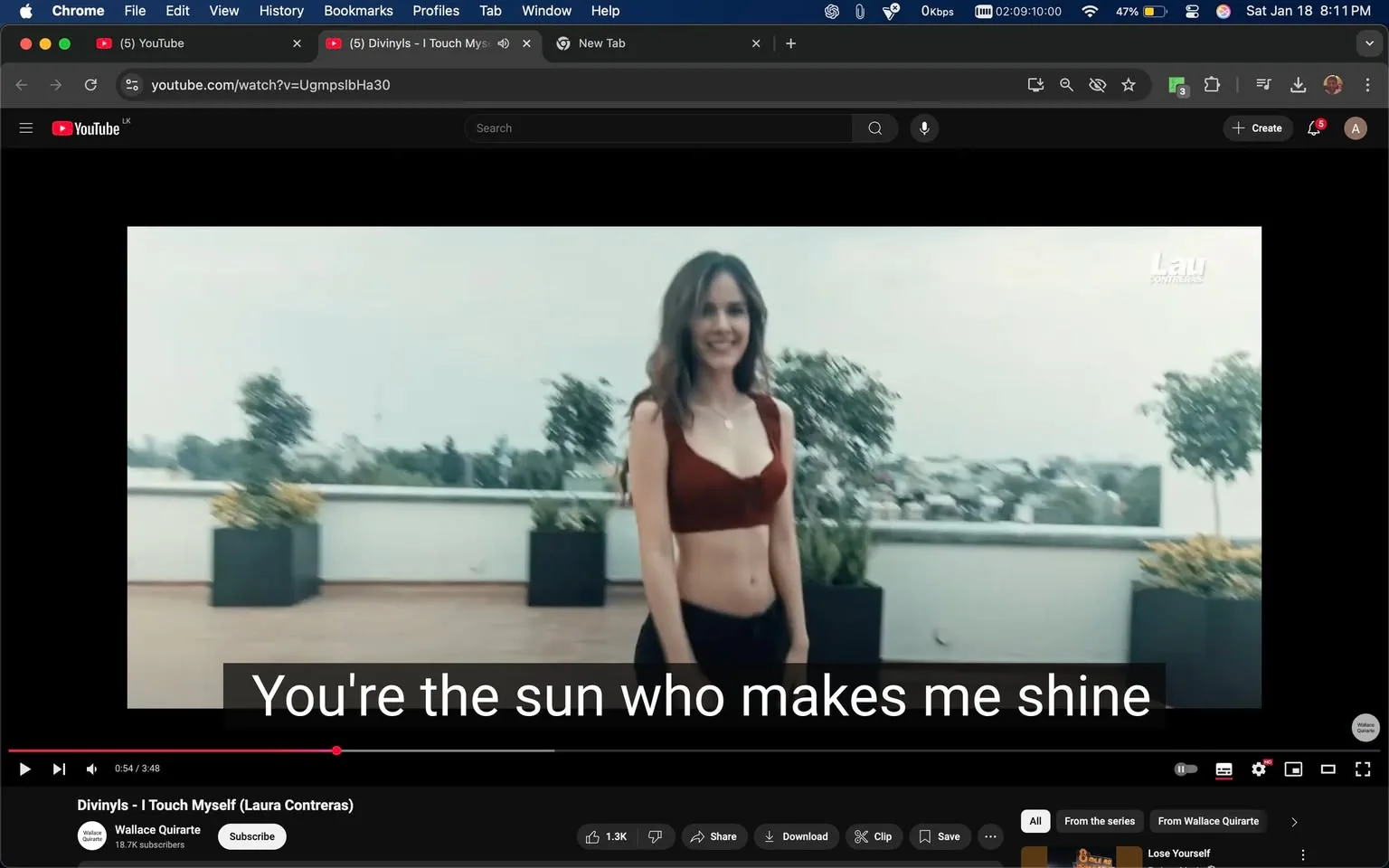 In the image, an unidentified adult woman stands on a rooftop terrace, smiling at the camera. She has long, wavy hair and is wearing a red crop top paired with black pants. The background features potted plants and a view of a cityscape, suggesting an outdoor setting. The atmosphere appears relaxed, and the lighting is soft, indicating it might be early evening. Below her, there are subtitles that read, "You're the sun who makes me shine," indicating that this scene is part of a music video. The overall vibe is casual and cheerful, with no visible signs of violence or explicit content.