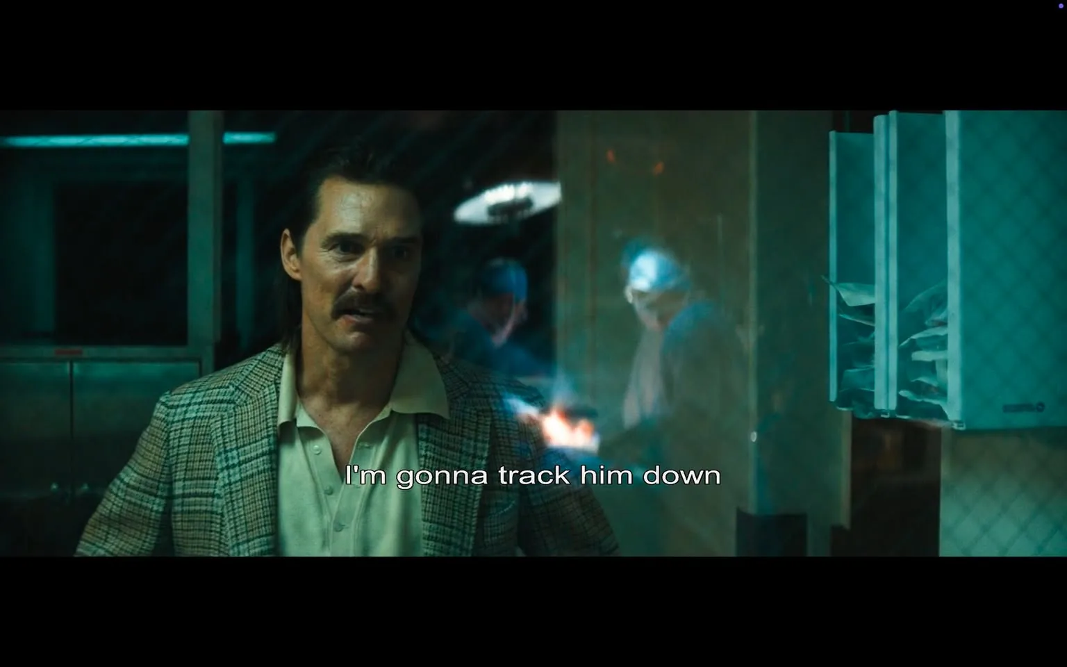 In the scene, a man with a prominent mustache and slicked-back hair is visibly agitated, declaring, "I'm gonna track him down." He wears a light-colored shirt under a patterned blazer, suggesting a retro or vintage style. The setting appears to be indoors, likely in a dimly lit room with a glass partition, hinting at a tense or serious atmosphere. Behind him, two figures are partially visible, engaged in an activity that seems to involve some form of work, possibly related to law enforcement or investigation, indicated by their attire and the context of the scene. The lighting casts a dramatic effect, enhancing the intensity of the moment. The presence of the glass partition and the characters' expressions suggest a narrative focused on pursuit or confrontation, with an underlying theme of conflict.