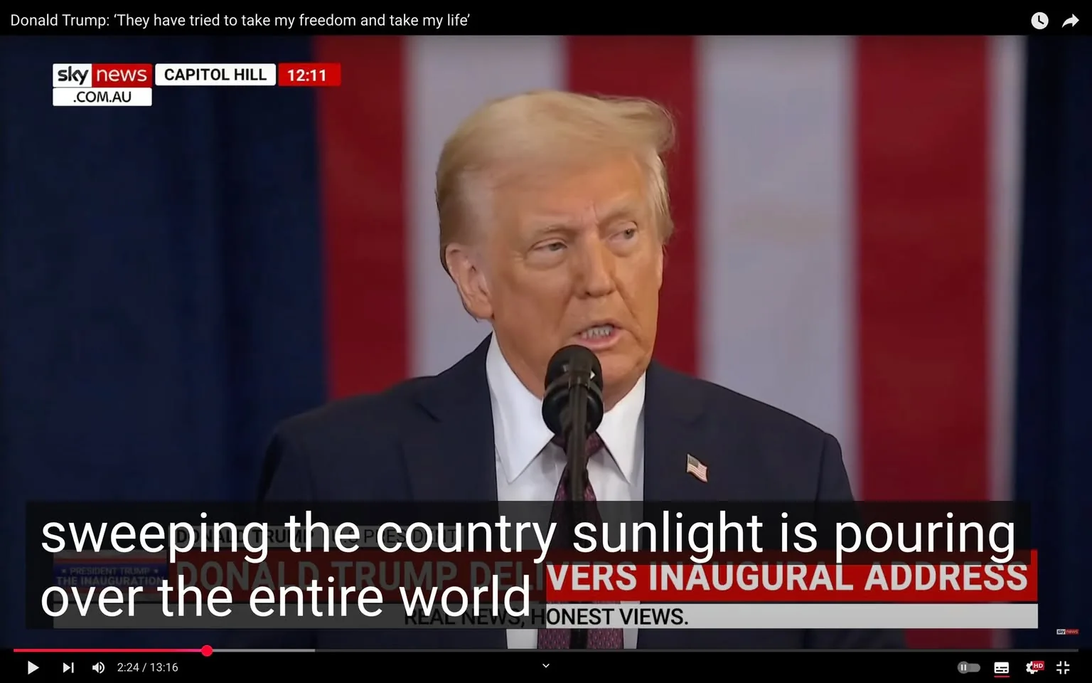 In the image, a public figure is seen speaking at a podium, delivering what appears to be an inaugural address. The individual has light-colored hair and is wearing a dark suit with a white shirt and a tie. The background features a large American flag, which suggests a patriotic setting. The text overlay indicates that this is a speech by Donald Trump, with a timestamp showing 12:11. The visible text includes a quote from the speech, discussing themes of freedom and personal struggle. The podium has a microphone, and the setting seems to be indoors, likely at a formal event. The branding for Sky News is visible in the upper left corner, indicating the source of the broadcast. Overall, the atmosphere is serious, reflecting the gravity of the speech being delivered.