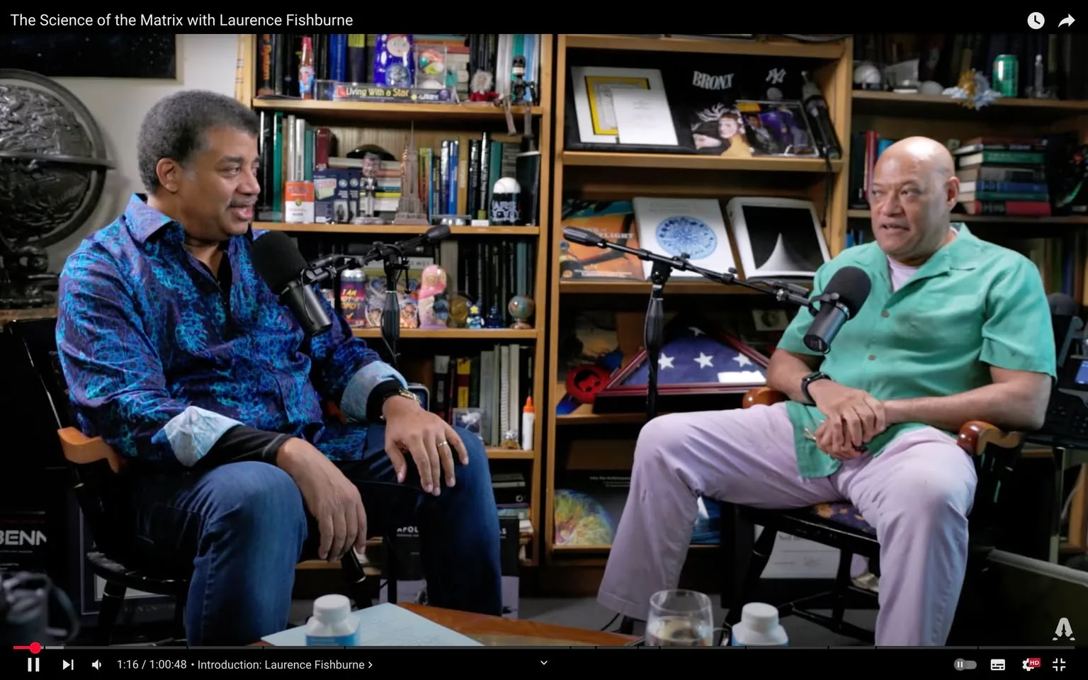 In a cozy indoor setting, two unidentified adults are seated across from each other, engaged in a conversation. One individual is wearing a vibrant blue patterned shirt with a black collar, while the other is dressed in a light green short-sleeved shirt paired with pale pink pants. Both are positioned in front of microphones, suggesting a recorded discussion or interview format. The background features a well-organized bookshelf filled with books, decorative items, and framed pictures, creating an intellectual atmosphere. There are also various objects on a table in front of them, including glasses and a bottle, indicating a casual yet professional setting. The overall ambiance suggests a focus on science and culture, likely related to the themes of their discussion.