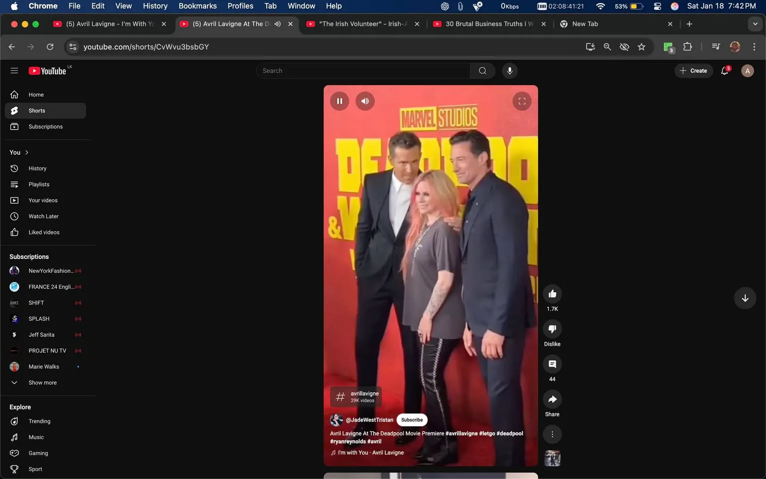 At a promotional event, three individuals are posing together in front of a vibrant backdrop featuring the text "DEADPOOL & WOLVERINE." The backdrop is predominantly red with yellow lettering, indicating a connection to the Marvel franchise. One individual, wearing a loose gray shirt and black pants with side zippers, has pink hair and stands in the center. The other two individuals are dressed in formal attire, one in a black suit and the other in a dark blue suit. They appear to be smiling and engaged with each other, suggesting a friendly atmosphere. The setting is likely indoors, given the controlled lighting and backdrop design typical of movie premieres. No sensitive content is present in the image.