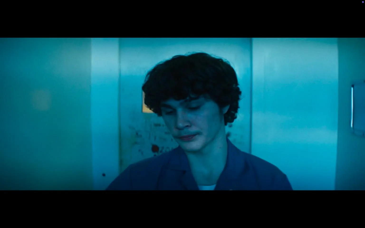 In the image, a young man with curly hair appears to be deep in thought or contemplation, standing in what looks like an elevator. The lighting is predominantly blue, creating a moody atmosphere. His expression is serious, with his gaze directed downward, suggesting a sense of introspection or concern. He is wearing a blue shirt, possibly a uniform, which adds to the somber tone of the scene. The background features walls that are somewhat worn, with visible marks and scratches, indicating that the elevator has seen considerable use. There are no visible logos or brands in the image, and the overall setting feels confined and somewhat claustrophobic. The absence of any other people or objects in the frame emphasizes the young man's solitude and emotional state.