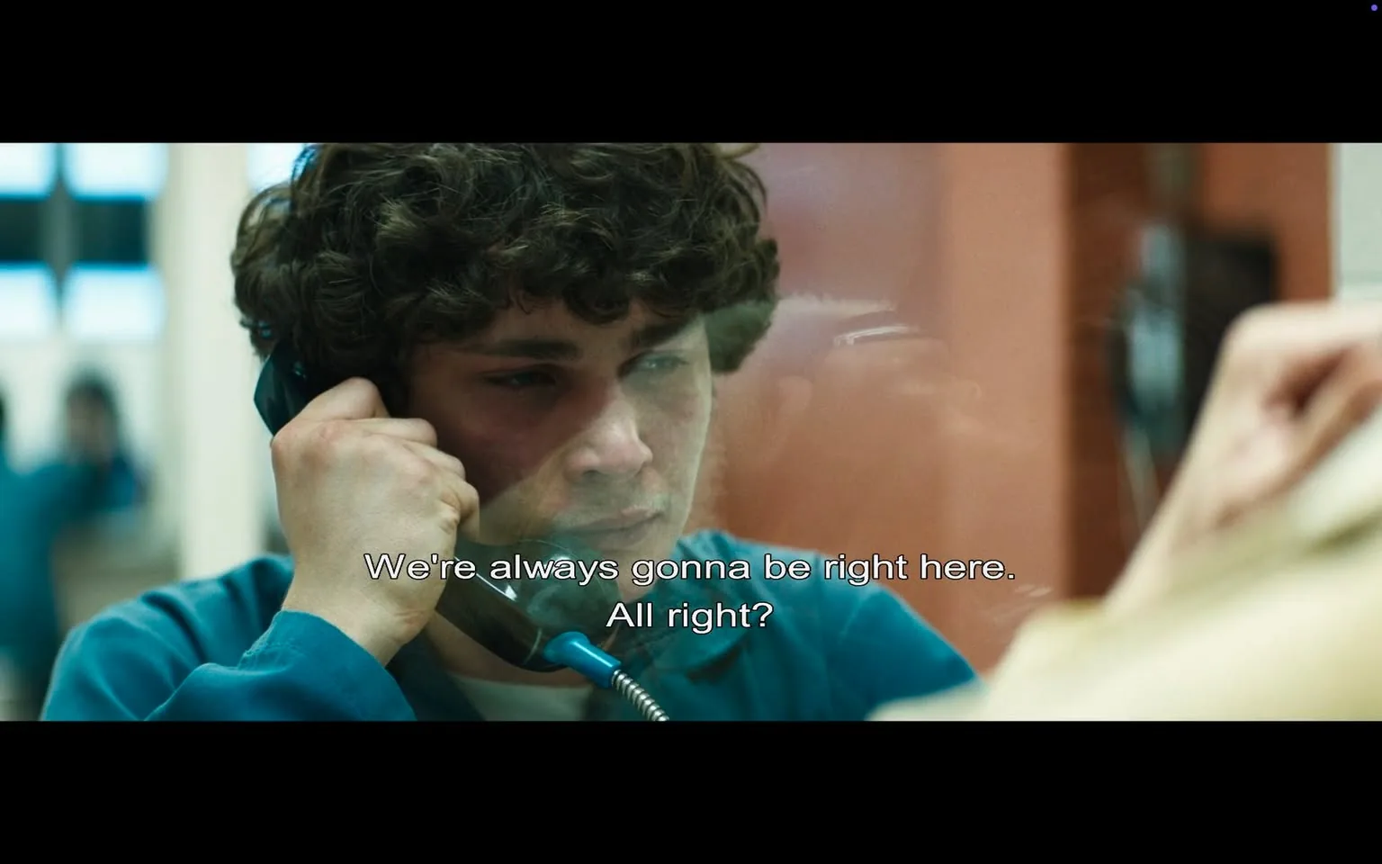 In the scene, a young man with curly hair is speaking on a telephone, appearing to be in a tense or emotional moment. He is wearing a blue prison uniform, which suggests he is in a correctional facility. His expression is serious and somewhat distressed, indicating the weight of the conversation. The background features blurred figures, likely other individuals in a similar setting, enhancing the atmosphere of confinement. 

The dialogue displayed on the screen reads, "We're always gonna be right here. All right?" This suggests a supportive or reassuring conversation, possibly between friends or family, indicating a bond despite the circumstances. The setting appears to be indoors, with a glass partition separating the young man from the person he is speaking to, which is a common feature in prison visitation areas. The overall mood is somber, reflecting themes of connection and struggle.