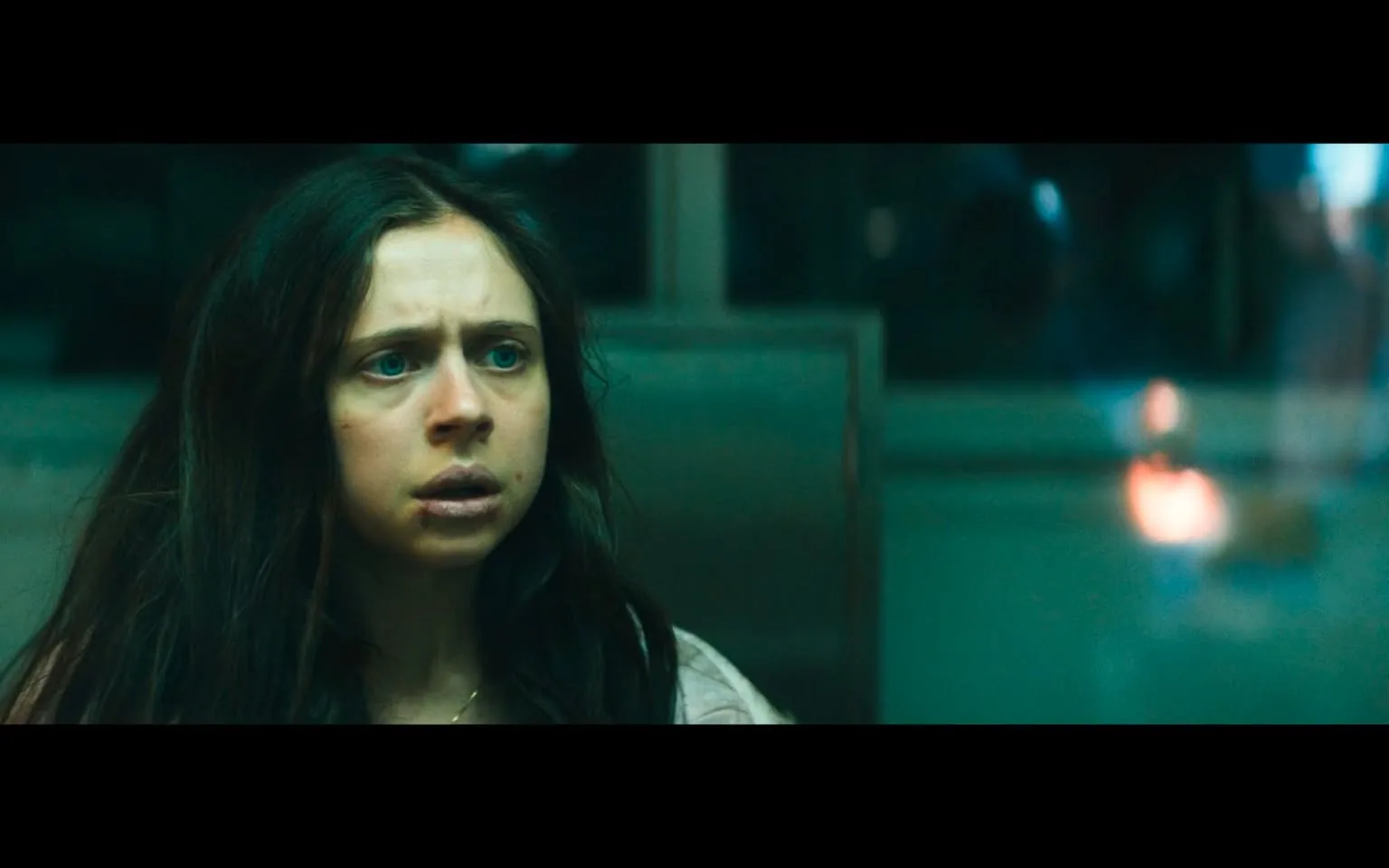 In the image, a young woman is seated, her expression one of fear and distress. She has long, dark hair that appears unkempt, and her skin looks pale, suggesting she may be unwell or in a state of shock. Her eyes are wide and a striking blue, conveying a sense of urgency or panic. There are visible signs of injury on her face, including what appears to be a bruise or cut on her lip, which adds to the impression of her being in a troubling situation.

The background is somewhat blurred, but it suggests an indoor setting, possibly a sterile or clinical environment, with muted colors that enhance the somber mood of the scene. The lighting is dim, contributing to the overall feeling of tension. The woman is wearing a simple, light-colored top, which looks slightly wrinkled, indicating she may have been in this situation for some time.

Overall, the image evokes a strong emotional response, hinting at themes of violence or trauma, as the woman's expression and injuries suggest she has endured a difficult experience.