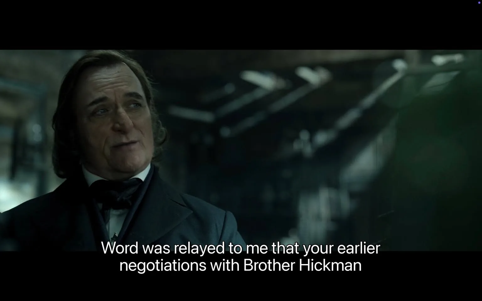 In the scene, a man with medium-length, wavy hair and a slight smile is engaged in conversation. He is dressed in a formal black suit with a white shirt and a black bow tie, suggesting a historical or period setting. The background appears dimly lit, with beams of light filtering through what seems to be an industrial space, hinting at an atmosphere of tension or secrecy. The man is speaking about previous negotiations with someone named Brother Hickman, indicating a serious discussion. His expression is calm yet slightly amused, as if he is reflecting on the absurdity of the situation. There are no visible logos, brands, or any explicit content in the image, maintaining a focus on the dialogue and the character's demeanor.