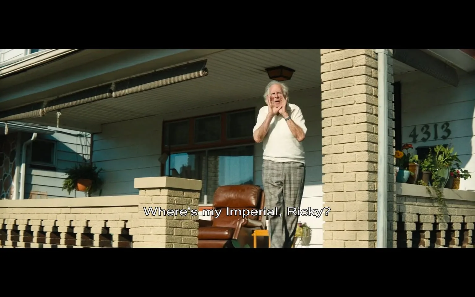 An elderly man stands on the porch of a house, visibly distressed as he raises his hands to his face in a gesture of concern or frustration. He is wearing a white short-sleeved shirt and plaid pants, suggesting a casual, homey attire. The setting appears to be a suburban neighborhood, with a light blue house in the background and a stone column supporting the porch. The porch features a brown leather chair and several potted plants, indicating a well-maintained outdoor space. 

The man is calling out, asking, "Where's my Imperial, Ricky?" This suggests a personal relationship with someone named Ricky, possibly a family member or friend. The time of day seems to be bright, likely daytime, as sunlight casts shadows on the porch. There are no visible signs of violence, sexual content, or any brands in the image. The overall atmosphere conveys a sense of urgency and concern regarding the missing item.