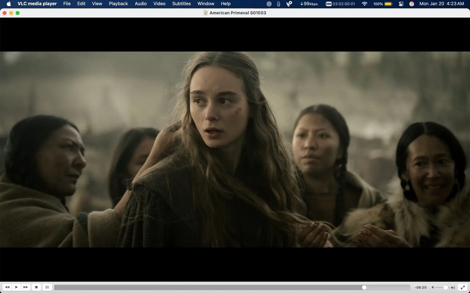 In the scene, a young woman with long, wavy hair appears to be the focal point, displaying a concerned expression as she looks to her left. She is dressed in a dark, textured garment, suggesting a historical or period setting. Surrounding her are three other women, each with distinct expressions; one appears to be comforting her, while another looks on with a neutral demeanor. The background is blurred, hinting at a rugged, outdoor environment, possibly a settlement or camp. The lighting is muted, contributing to a somber atmosphere, indicative of a tense or emotional moment. No visible brands or logos are present in the image.
