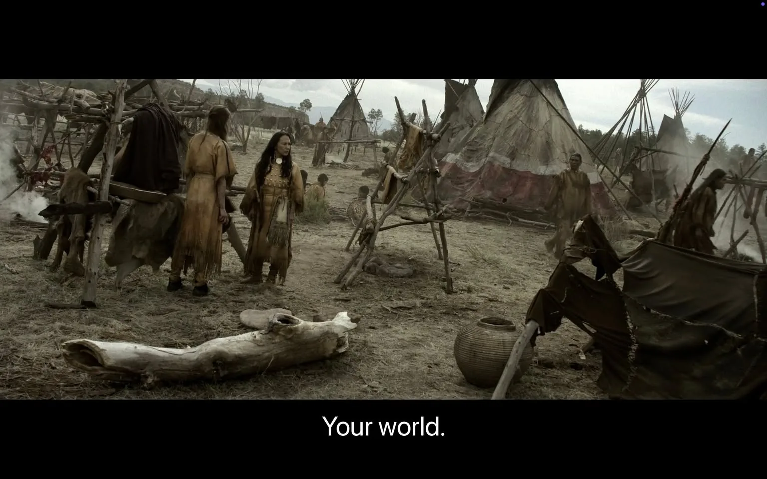 In a historical setting, a group of individuals dressed in traditional attire is present, suggesting a cultural or tribal context. The scene features several tents made of natural materials, indicating a communal living space. The individuals appear engaged in conversation, with one person gesturing, possibly emphasizing a point. The landscape is barren, with dry earth and sparse vegetation, contributing to a somber atmosphere. In the background, additional figures can be seen, some involved in various activities, while others are seated. The overall mood reflects a moment of interaction within a community, set against a muted, overcast sky.